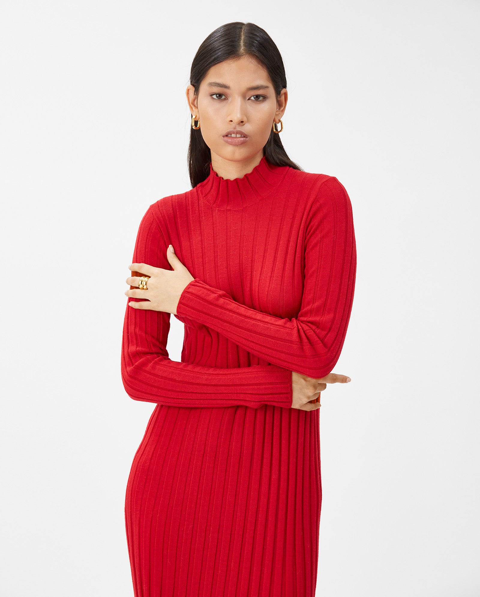 Woman with long dark hair wears the IVY OAK KAMELIA Dress, a fitted, ribbed red maxi knit, styled with gold hoop earrings and arms crossed.