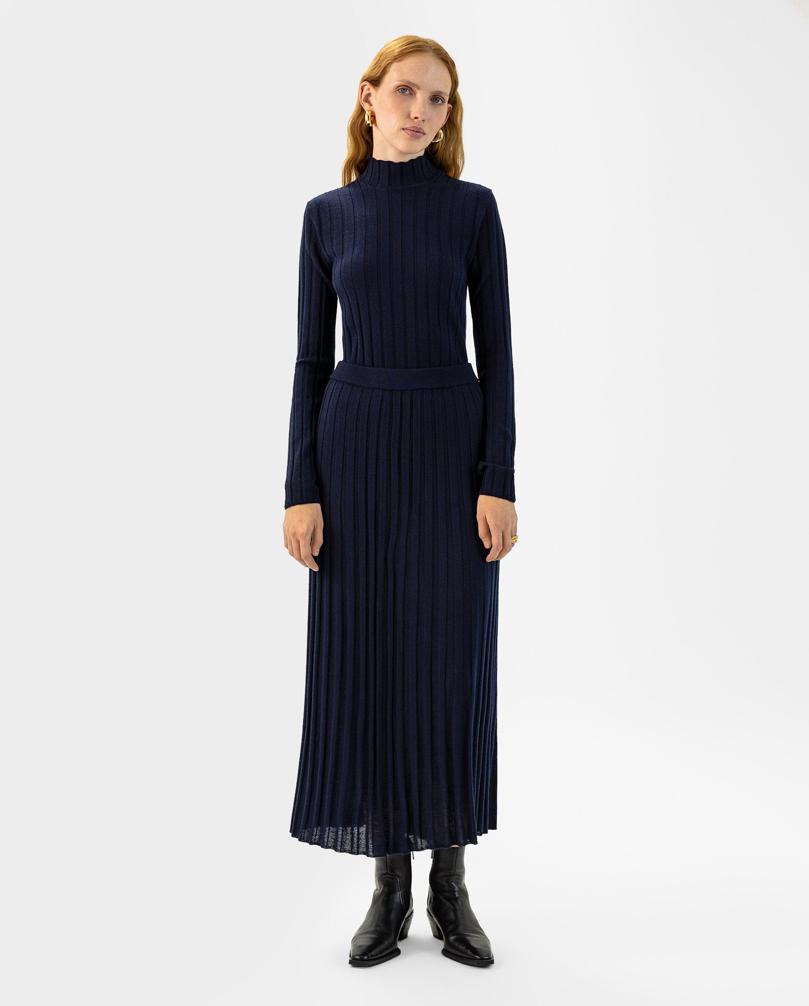 A woman wearing the IVY OAK KALI Skirt styled with a long-sleeved, navy ribbed knit dress featuring a flared skirt and high neck in soft merino wool, paired with black ankle boots.