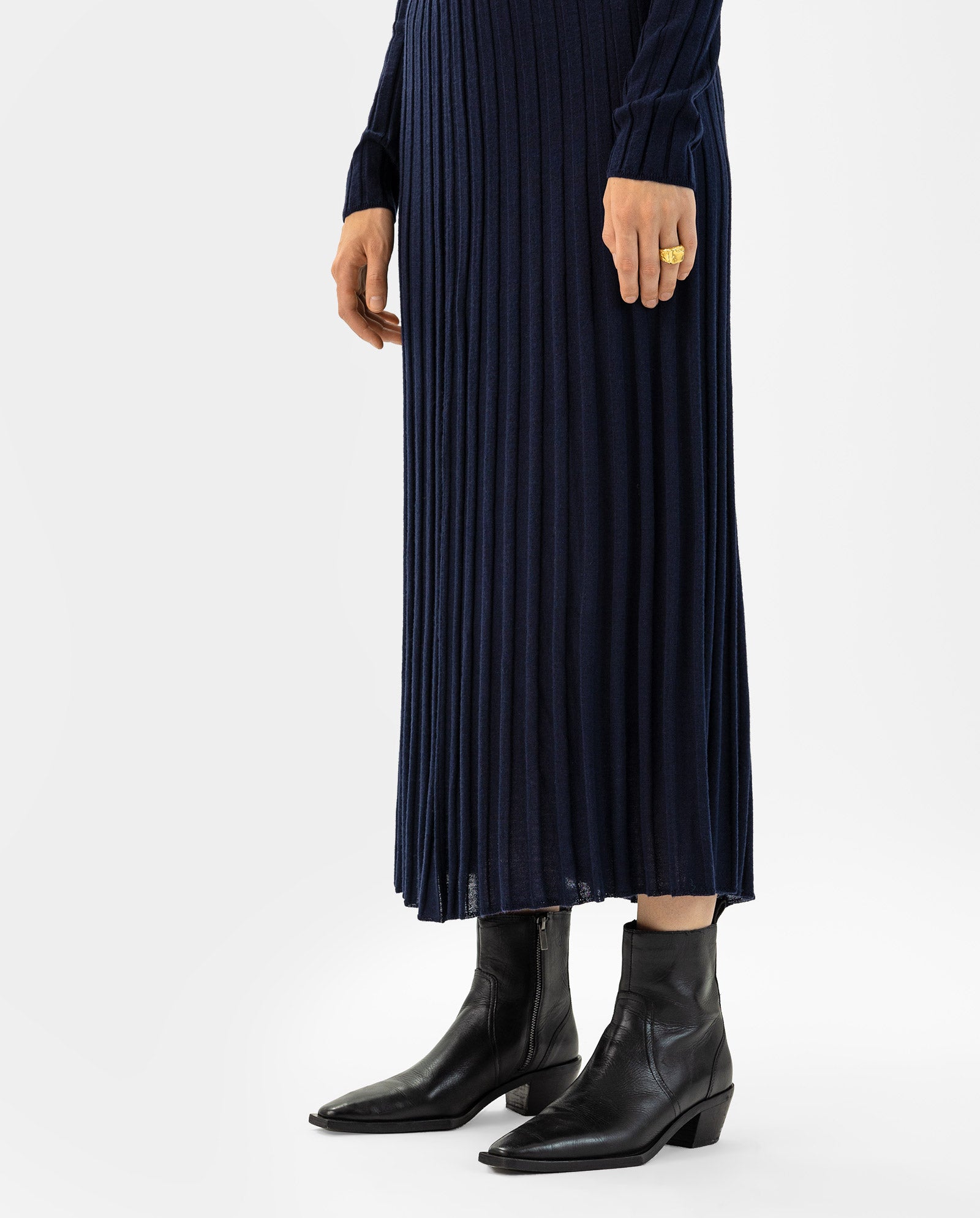 A person wears the IVY OAK KALI Skirt, a navy midi flared knit skirt in soft merino wool, paired with black ankle boots against a white background.
