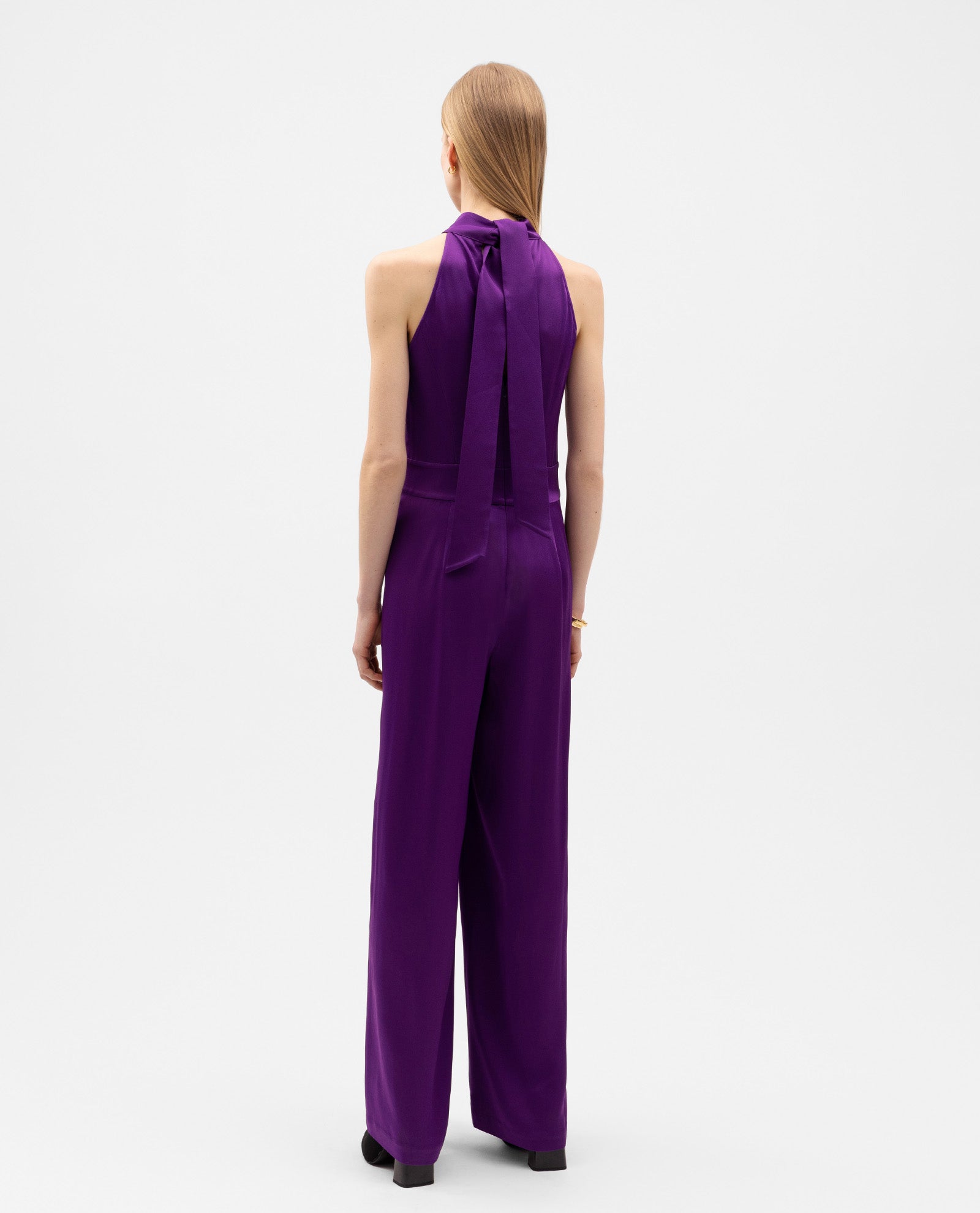 A person is seen from the back wearing the IVY OAK PHILIPPINE Jumpsuit—a sleeveless, purple satin design featuring wide legs and a halter-neck.