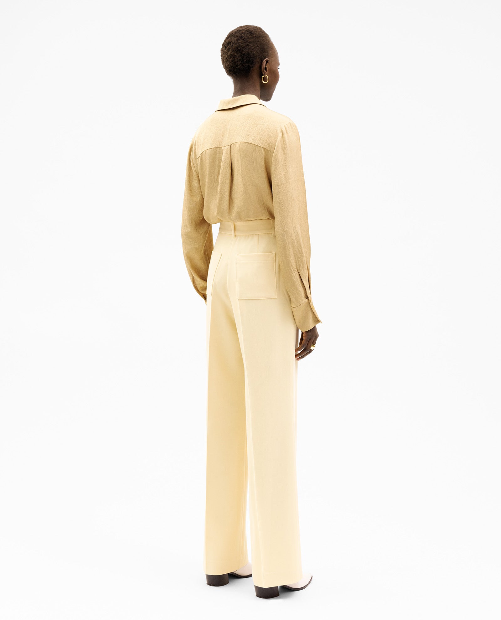 A person stands facing away, wearing a tan shirt, heeled boots, and the IVY OAK PENINA Trouser—cream high-waisted woven trousers with patched pockets.