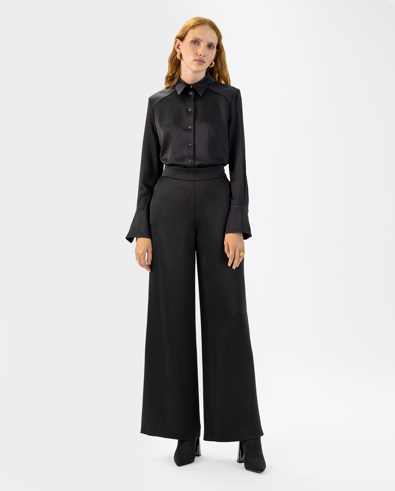 A woman in a black button-up shirt and IVY OAK’s PETUNIA Trouser, featuring a high waist and wide-leg silhouette, stands against a plain white background.