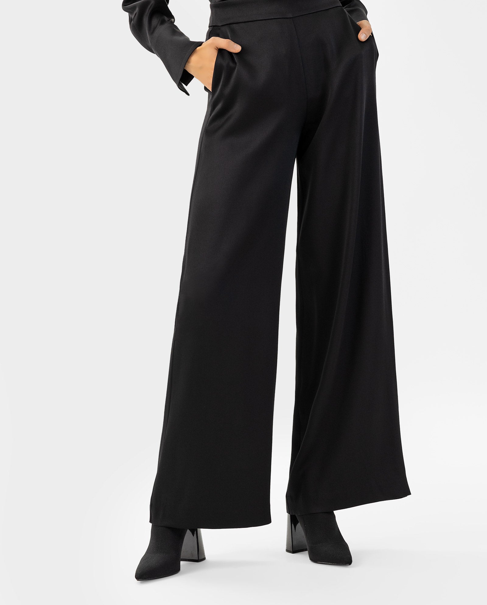 A person wears the IVY OAK PETUNIA Trouser—high-waisted, wide-leg black pants—with heeled boots and hands in pockets, set against a plain background.