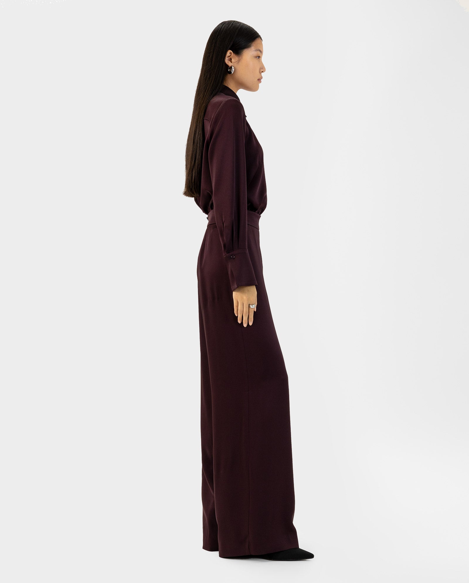 A woman stands sideways against a plain white background, wearing IVY OAKs PETUNIA Trouser—a long-sleeved, dark burgundy outfit with high-waisted pants.