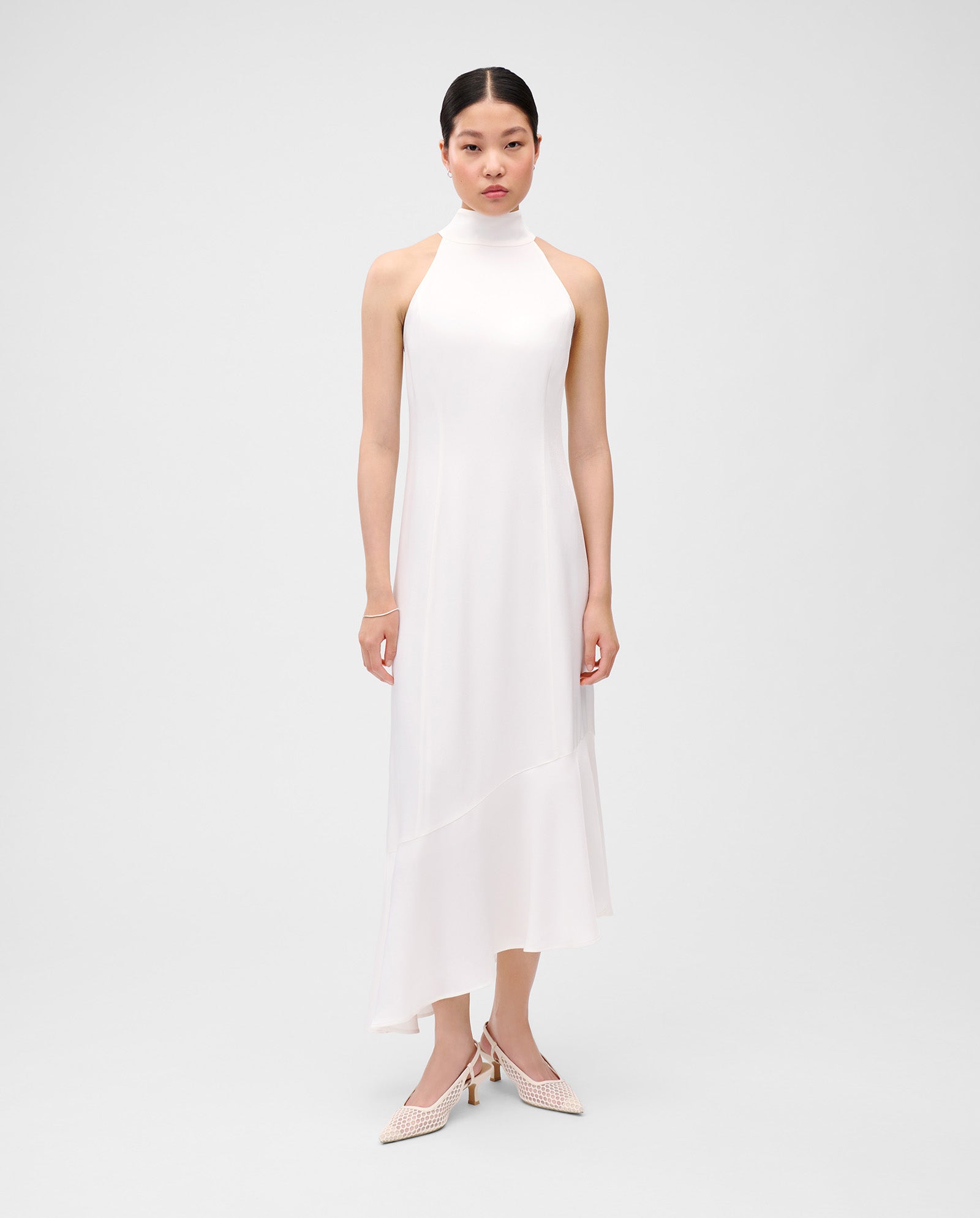 A woman in the NONA Dress by IVY OAK—a sleeveless, high-neck white satin dress with princess seams, asymmetrical hem, and concealed zip—stands against a plain background.