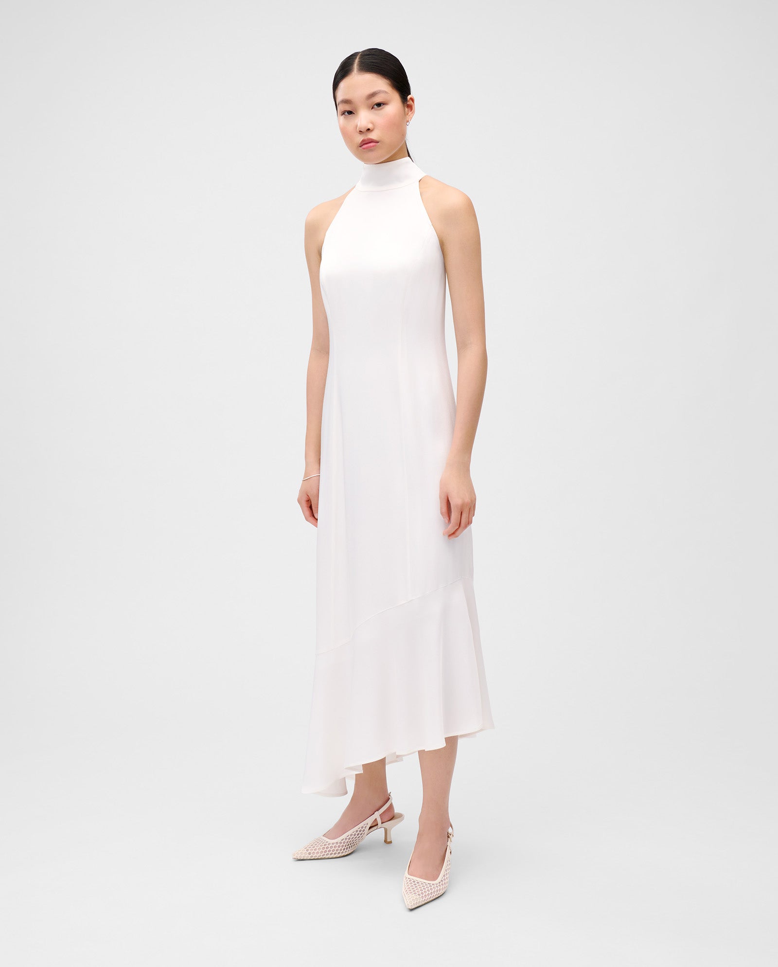 A woman wears the IVY OAK NONA Dress—a sleeveless, high-neck white satin dress with princess seams and a concealed zip—paired with pointed heels, against a plain background.