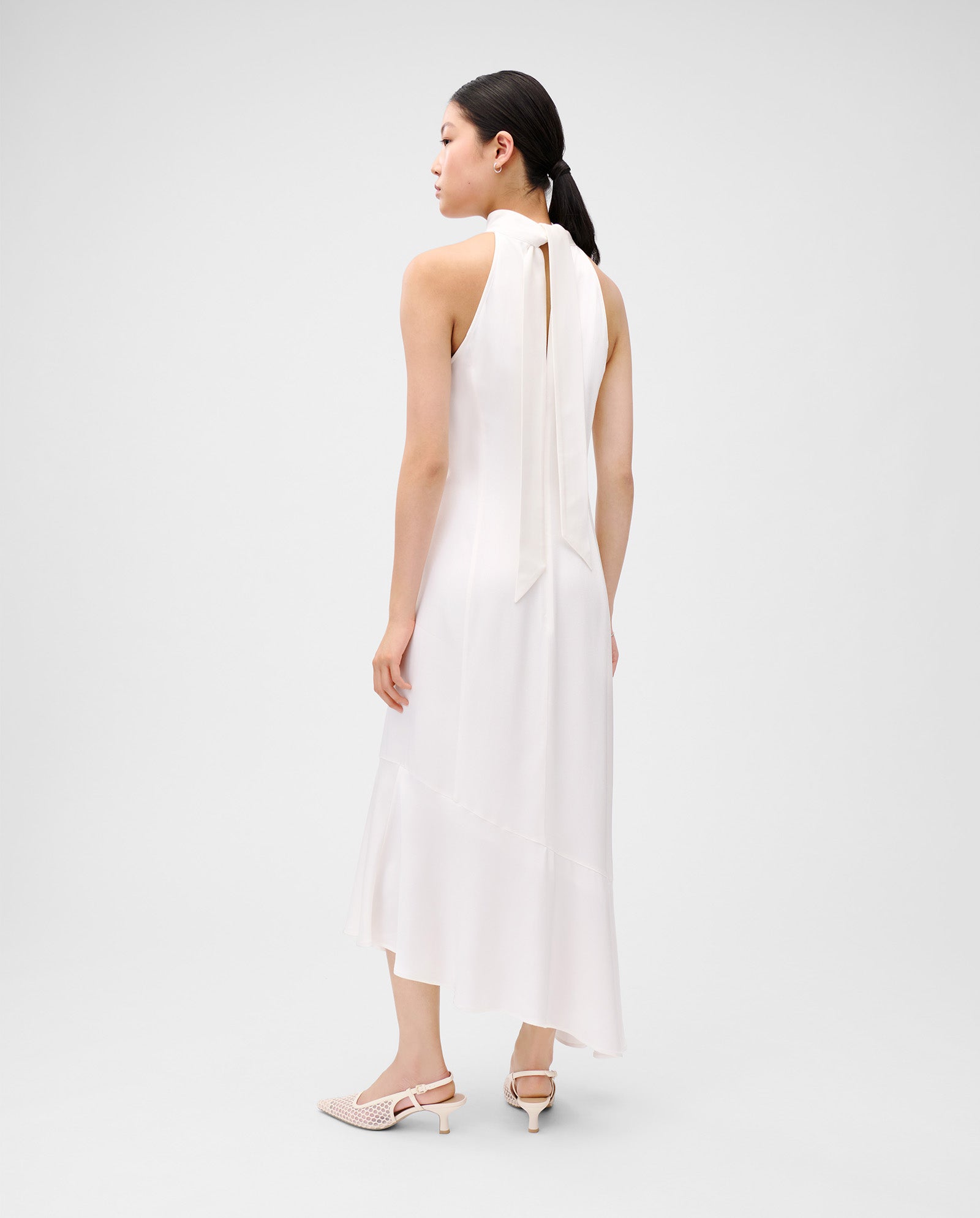 A woman wears the IVY OAK NONA Dress, a sleeveless light ivory satin piece with princess seams, tie-neck back, concealed zip, and white slingback heels.
