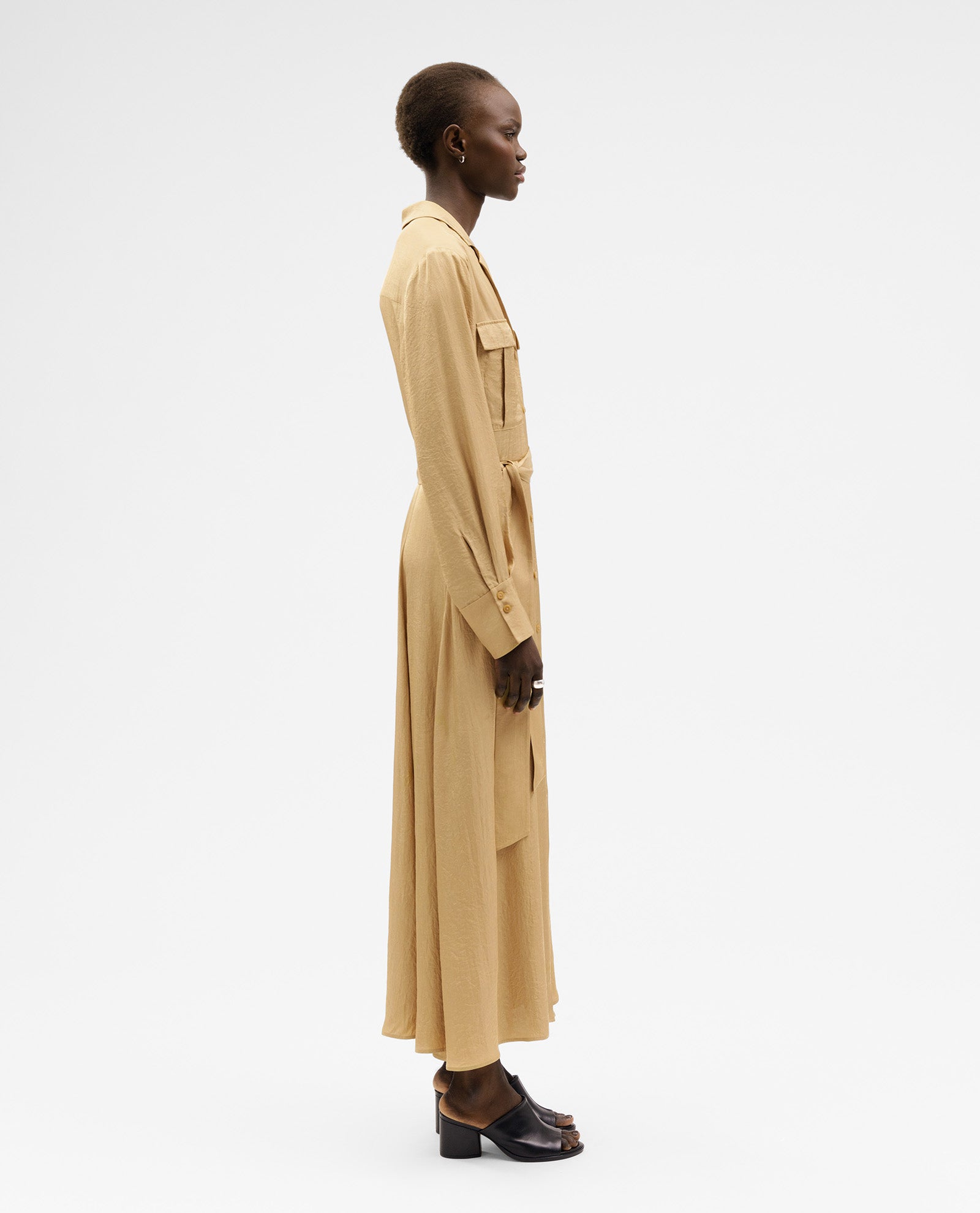 Woman in profile wears the IVY OAK NYLA Dress—a long beige maxi style with a belted waist and button placket—paired with black heeled sandals against a white background.