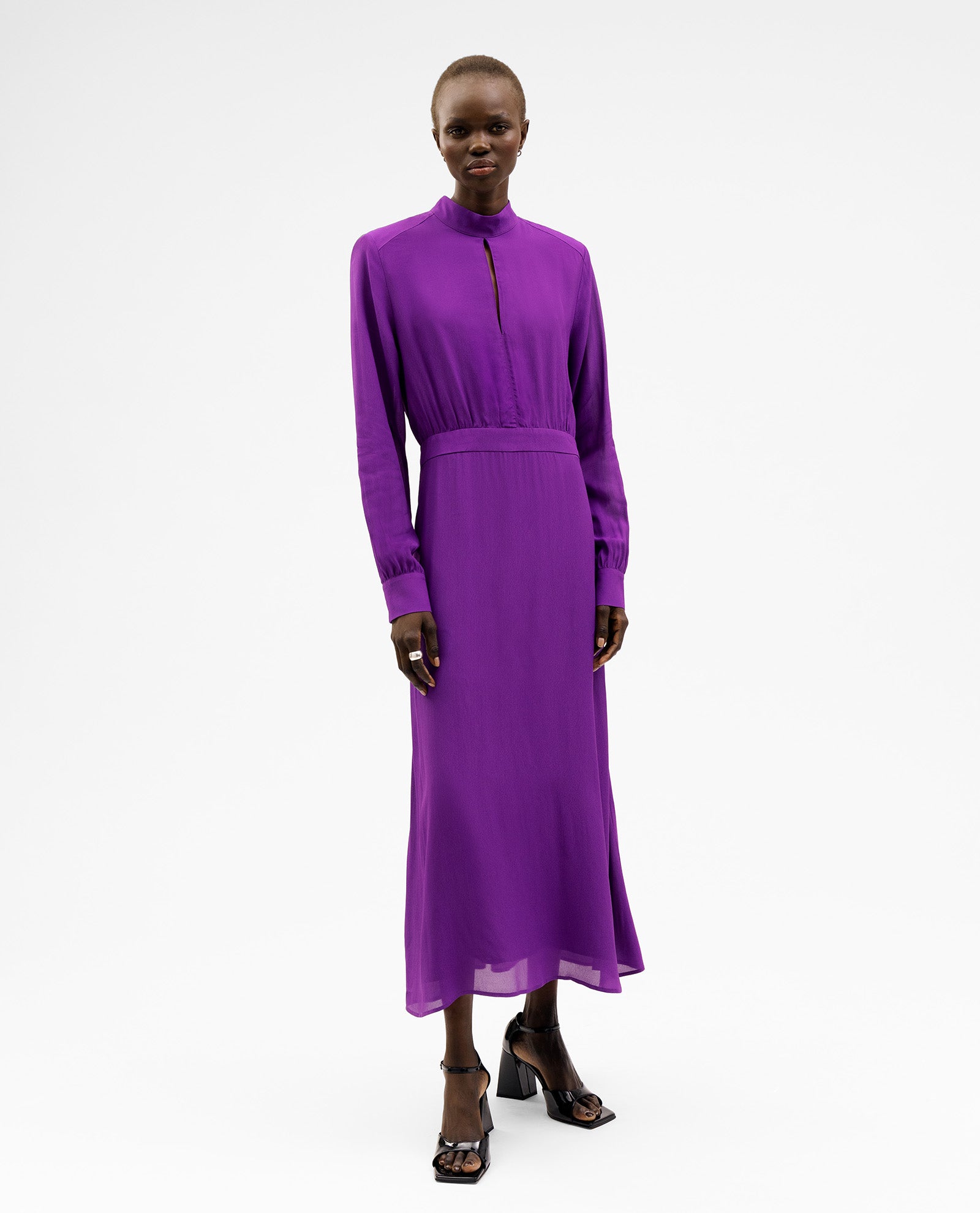 A woman wearing the long-sleeved, purple NEVE Dress by IVY OAK with Schulterpads and black heeled sandals stands against a white background.