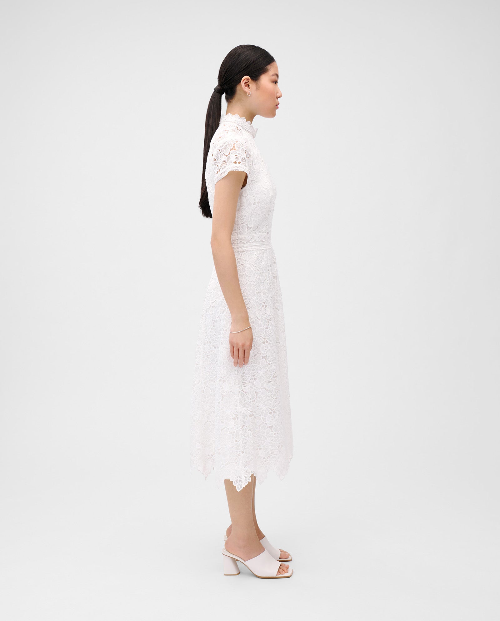 A woman in the IVY OAK NESSY Dress with floral lace and a flared fit, paired with heeled sandals, stands in profile against a plain background.