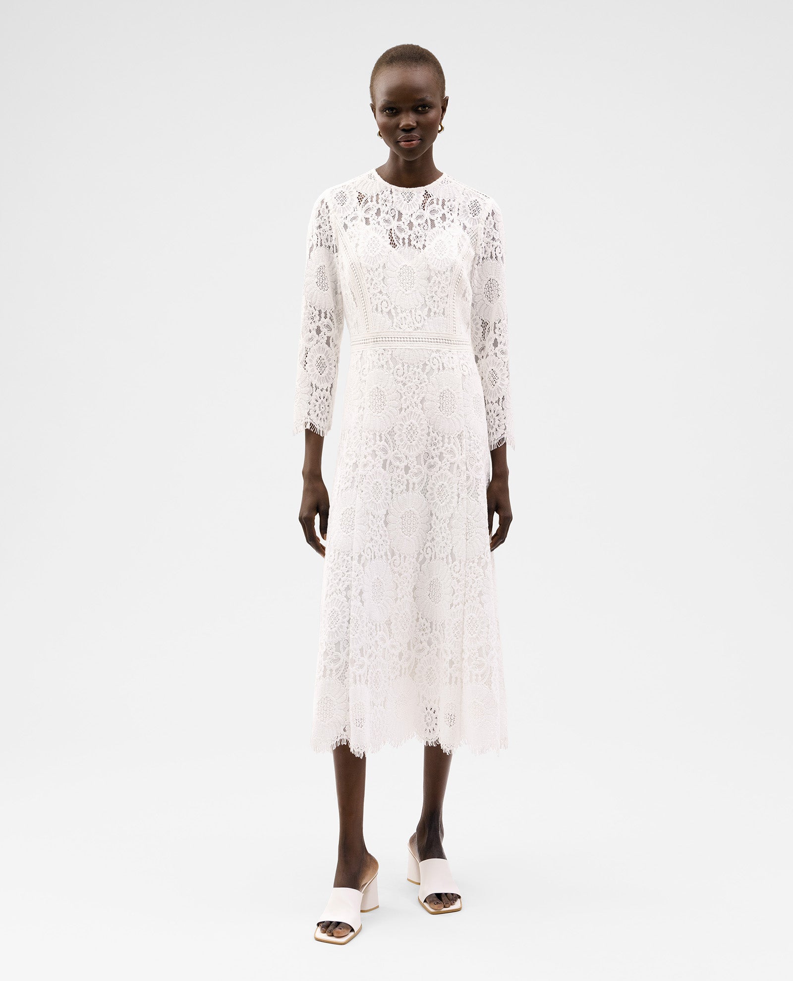 A woman stands against a plain background wearing the IVY OAK MAGDA Dress, a long-sleeve, midi floral lace bridal gown with a fitted zipper, paired with white open-toe heels.