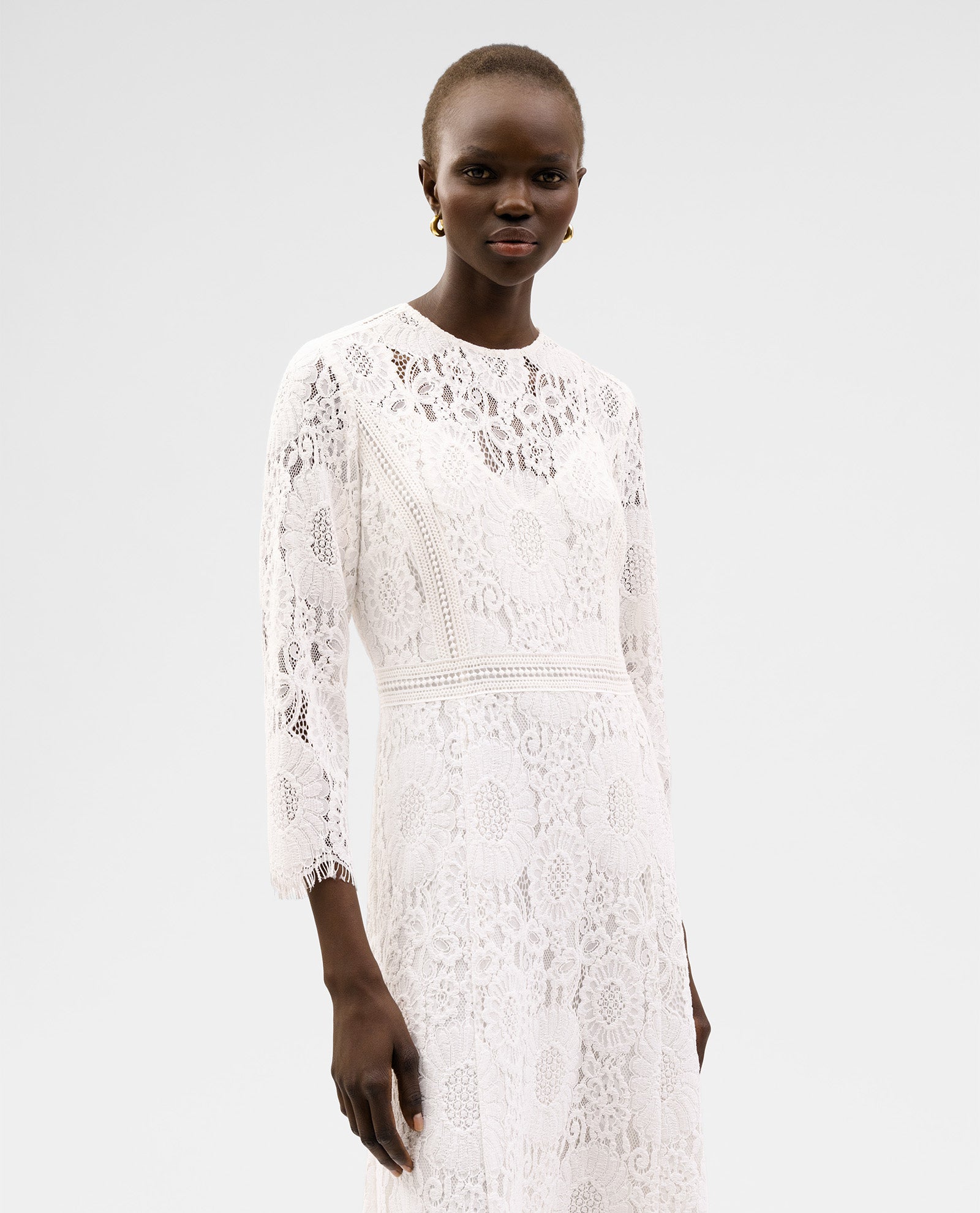 A woman wears the IVY OAK MAGDA Dress, a midi floral lace bridal gown featuring long sleeves and a fitted zipper, standing against a plain light background.