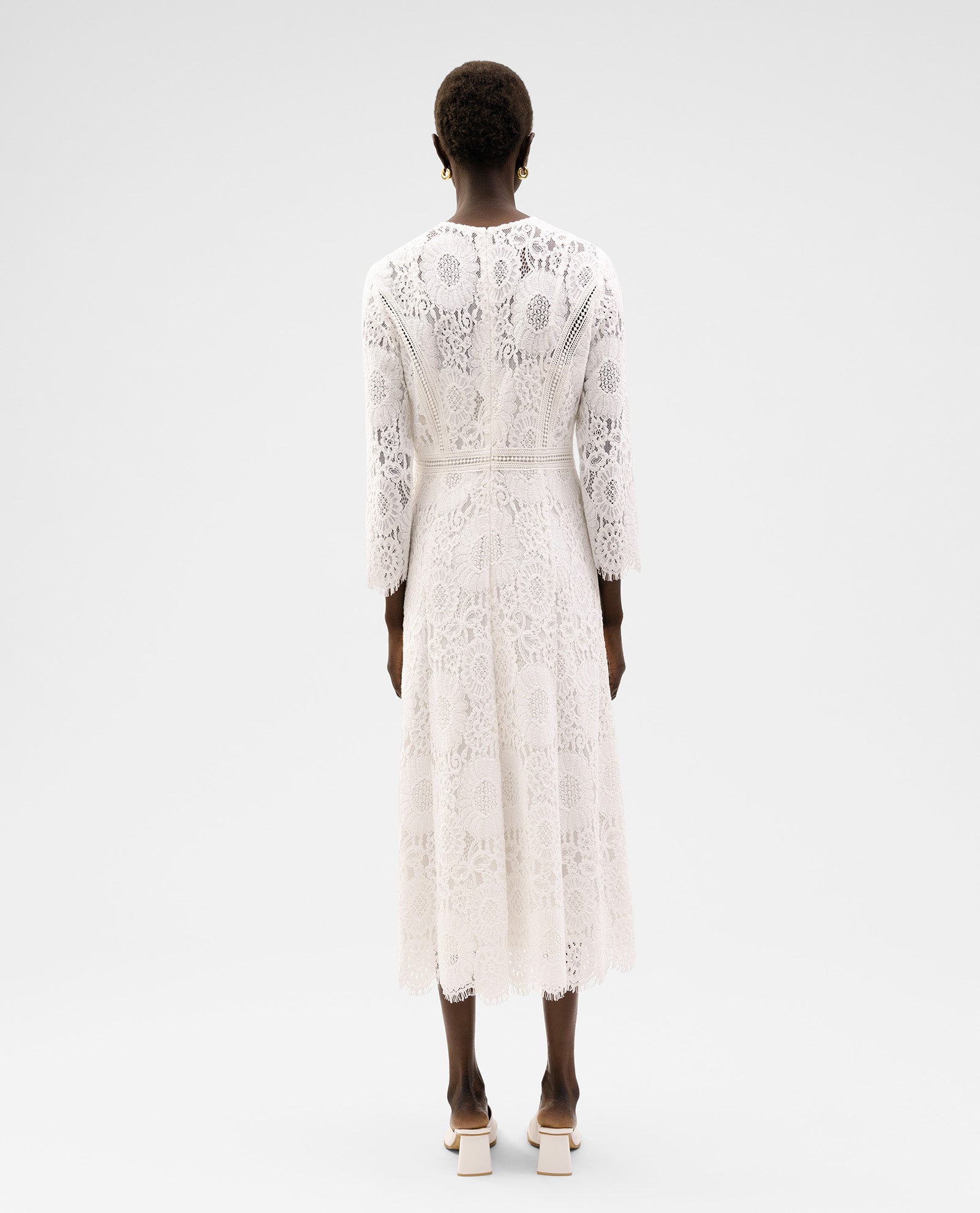 A person is seen from the back against a plain background wearing the IVY OAK MAGDA Dress, a long-sleeved midi floral lace piece with a fitted zipper, styled with heels.