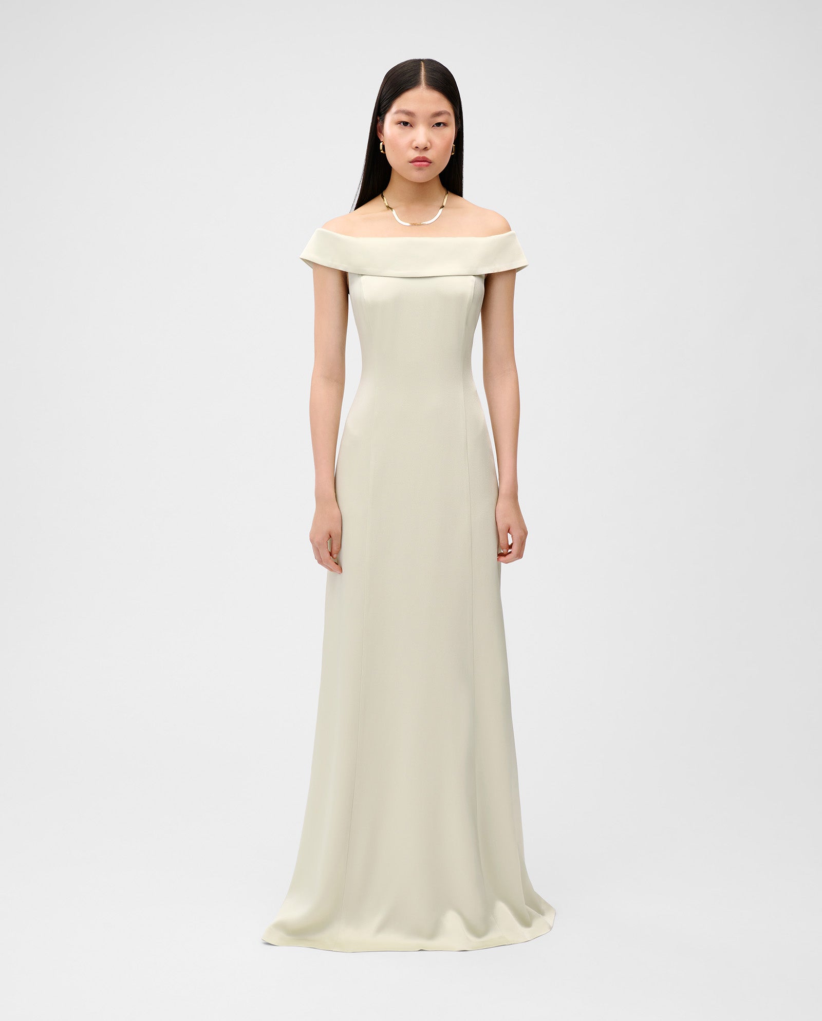 A woman wears the NADELLE Dress by IVY OAK in light beige, styled with a pearl necklace. The maxi off-shoulder dress features an elegant lining and subtle zipper, and she stands against a plain background.