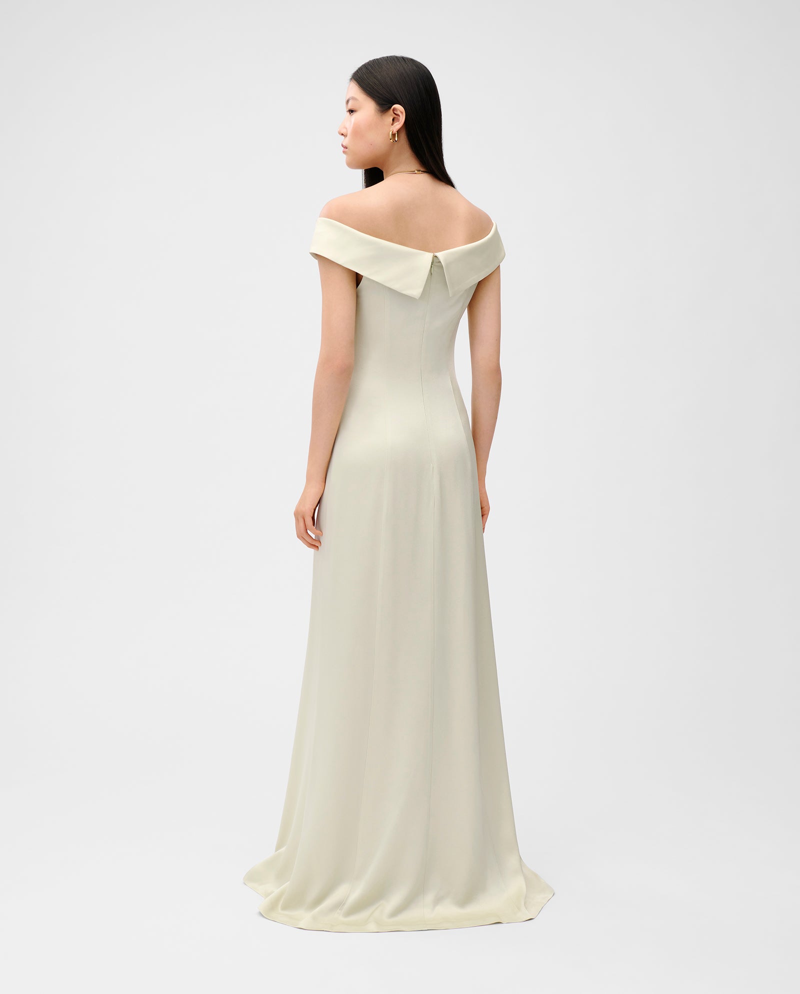 A woman wearing the NADELLE Maxi off-shoulder dress by IVY OAK stands facing away against a plain light background.