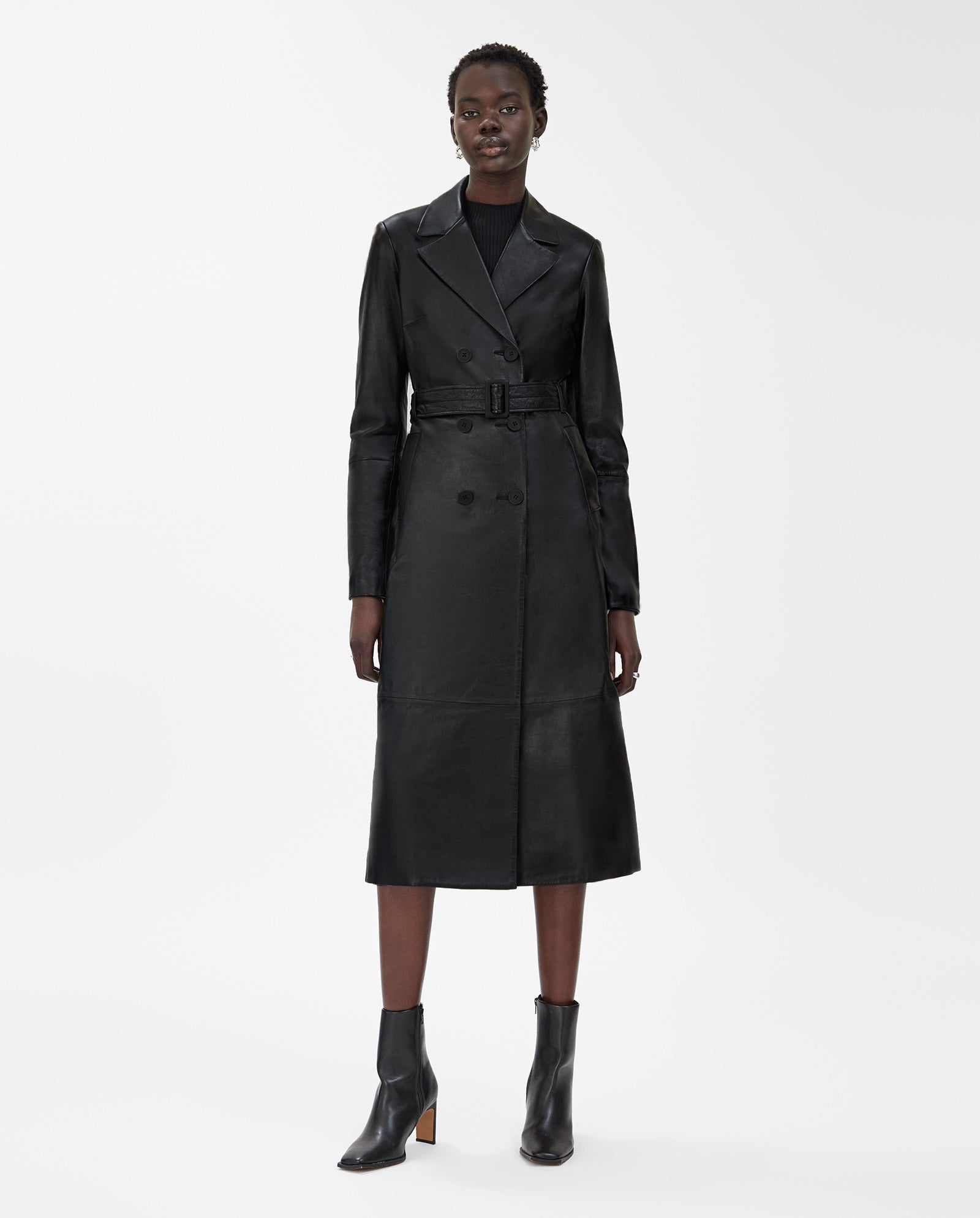 A woman wears the IVY OAK LIONNE midi black lamb nappa leather trench coat with a belt and ankle boots, standing against a plain white background.
