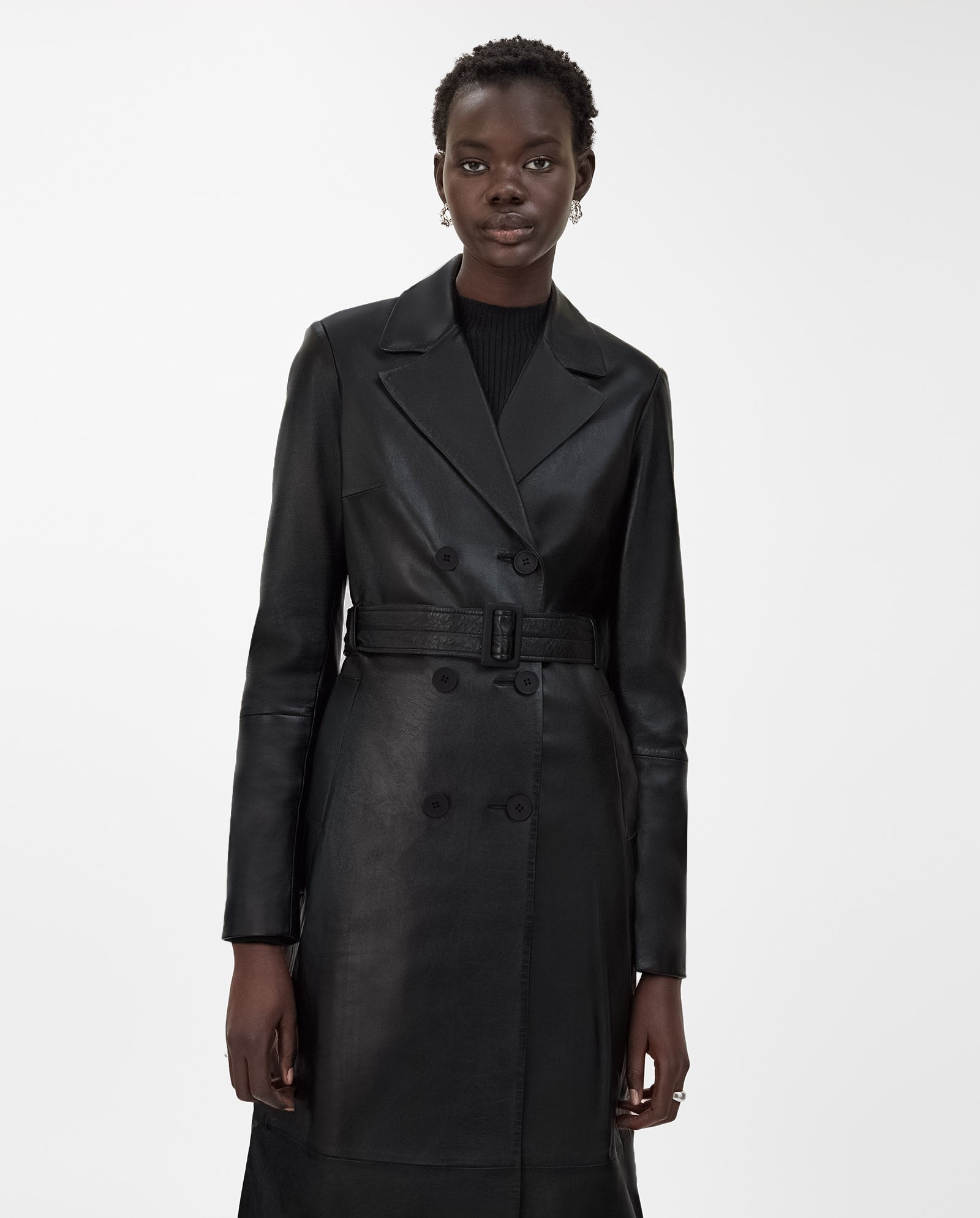 A woman stands against a plain white background wearing the IVY OAK LIONNE Coat, a belted, midi-length black lamb nappa leather trench, styled over a black top.