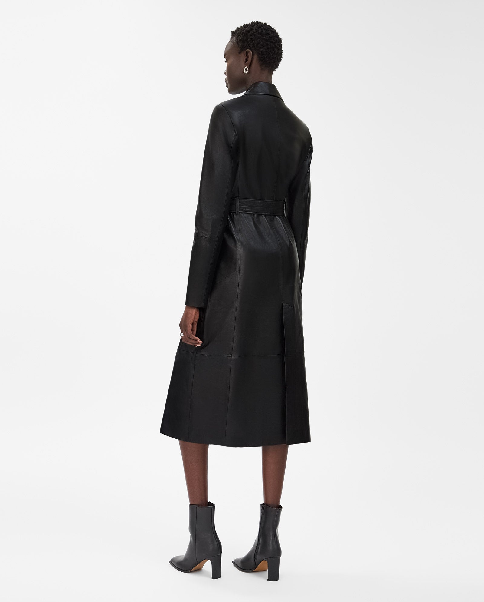 A woman stands facing away on a white background, wearing the IVY OAK LIONNE coat—a black, midi-length leather trench crafted from lamb nappa—paired with ankle boots.