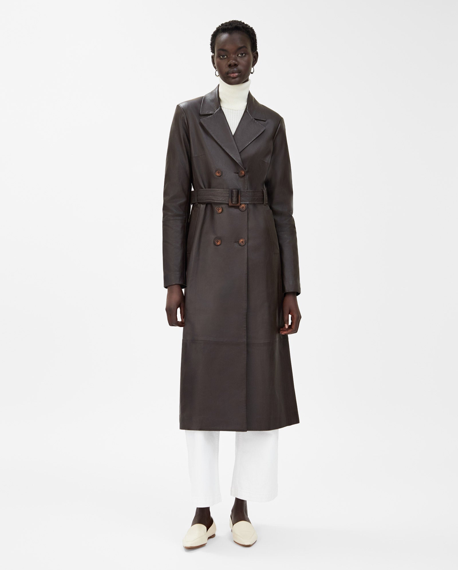 A woman wears the IVY OAK LIONNE long dark brown leather trench coat over a white turtleneck and white pants.