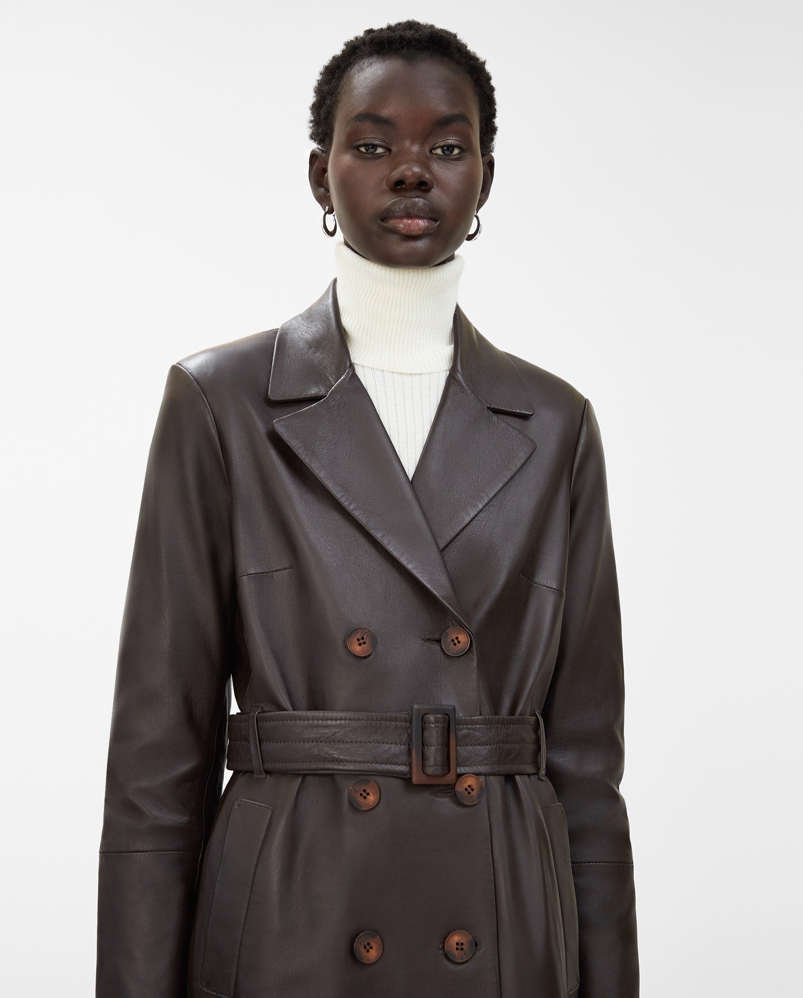 A woman stands against a plain background wearing the IVY OAK LIONNE coat, a dark brown midi-length trench crafted from lamb nappa leather, belted at the waist over a white turtleneck.