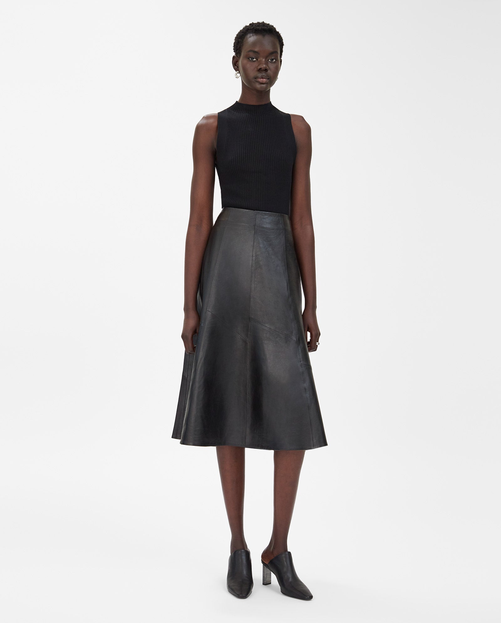 A woman in a sleeveless black top, the IVY OAK LAUREEN black leather flared midi skirt, and black heeled mules stands against a white background.