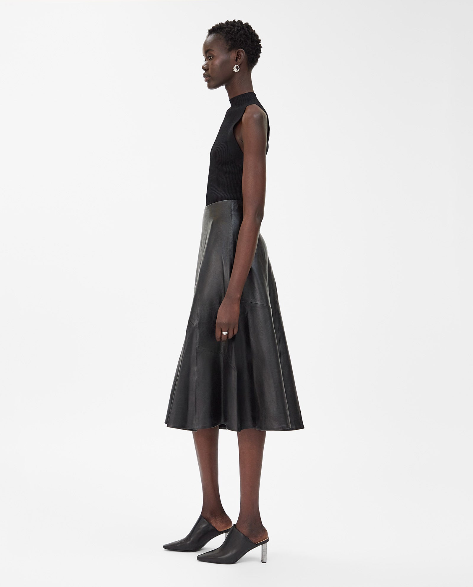 A woman in a sleeveless black top, IVY OAKs LAUREEN Skirt in flared-fit black leather, and black heeled mules stands in profile.