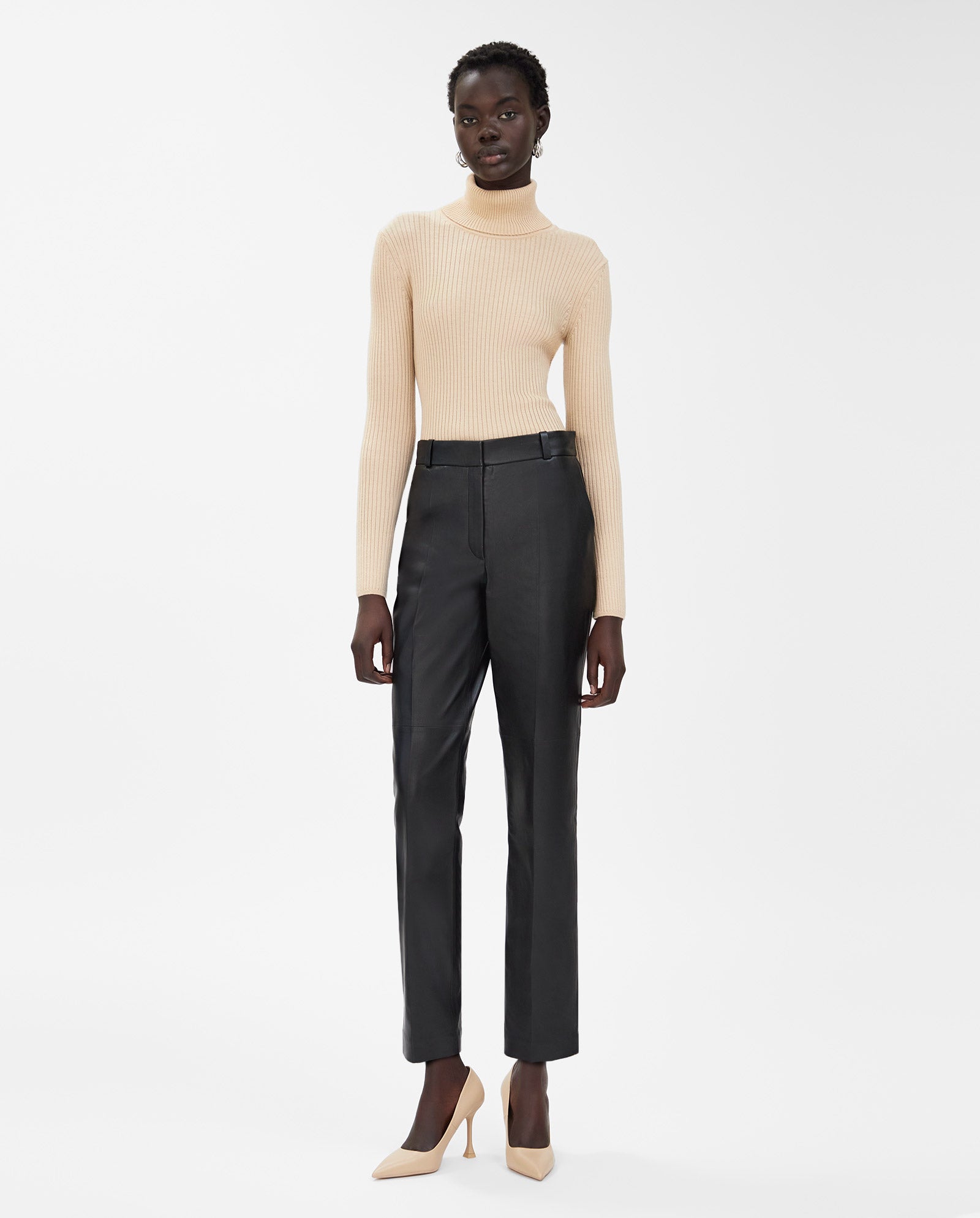 A woman wearing a beige turtleneck, IVY OAK LAURA slim fit high waist leather pants, and beige heels stands against a white background.