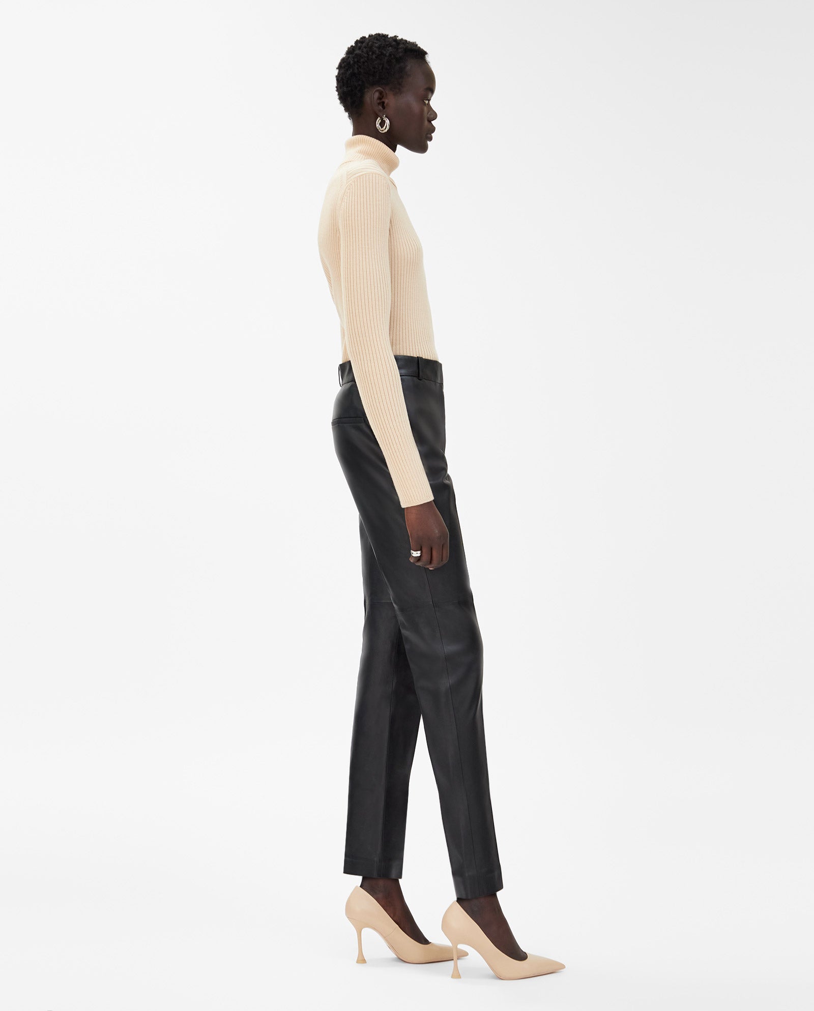 A person in a beige turtleneck, IVY OAK LAURA black leather slim-fit pants, and beige heels stands in profile against a white background.