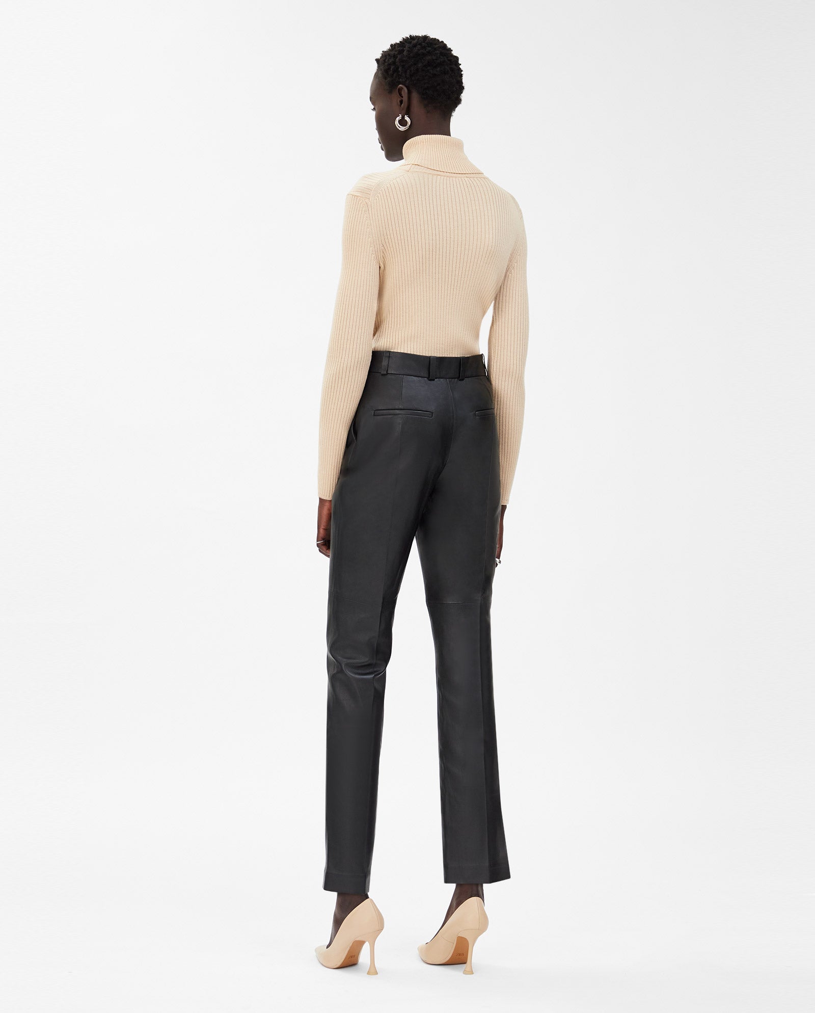 Woman standing in IVY OAK’s LAURA Pants—high waist black leather trousers—paired with a beige turtleneck and beige heels, facing away from the camera.