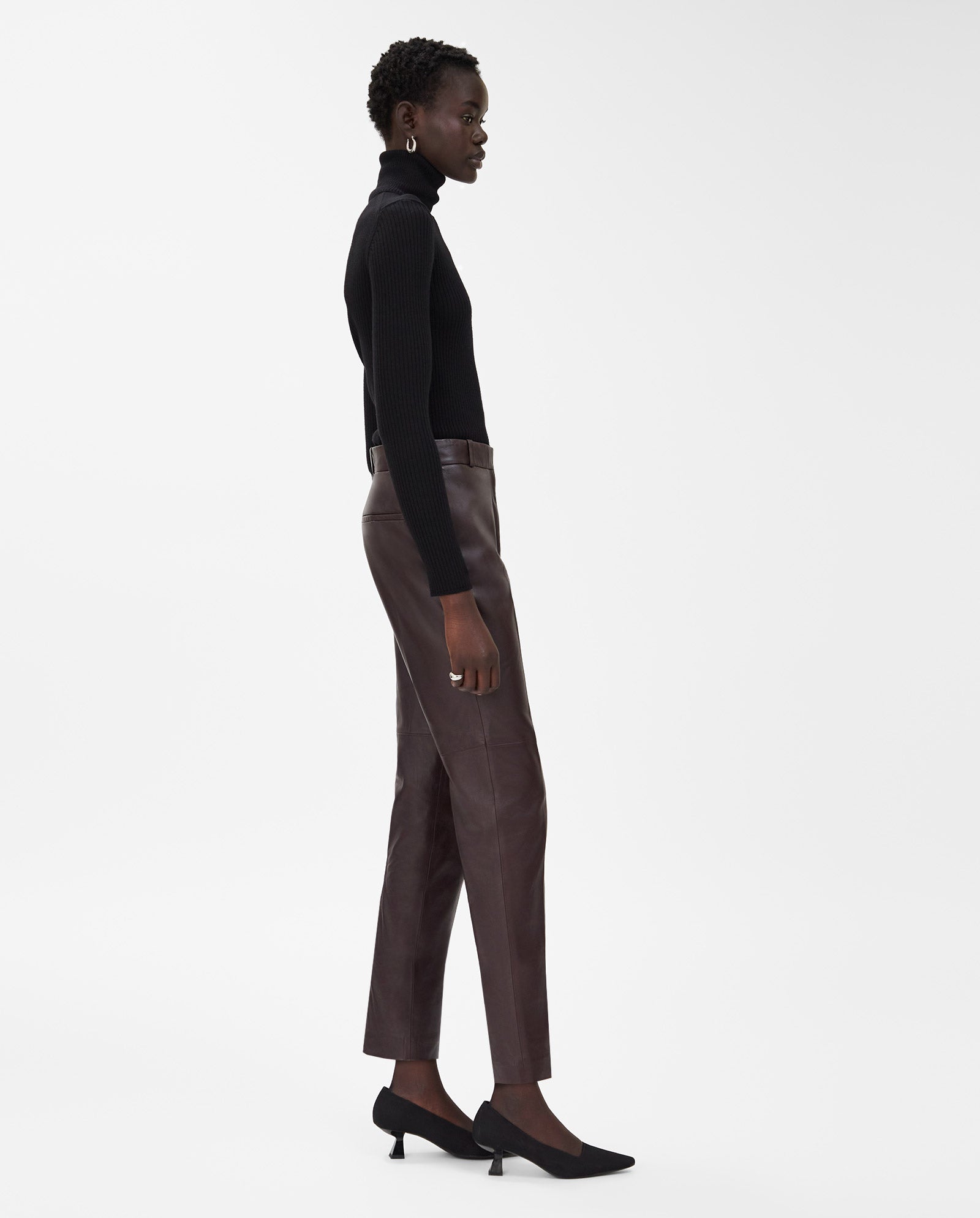 A woman stands in profile against a white background, wearing IVY OAKs LAURA Pants—sleek black slim-fit leather trousers—paired with a black turtleneck and black heels.