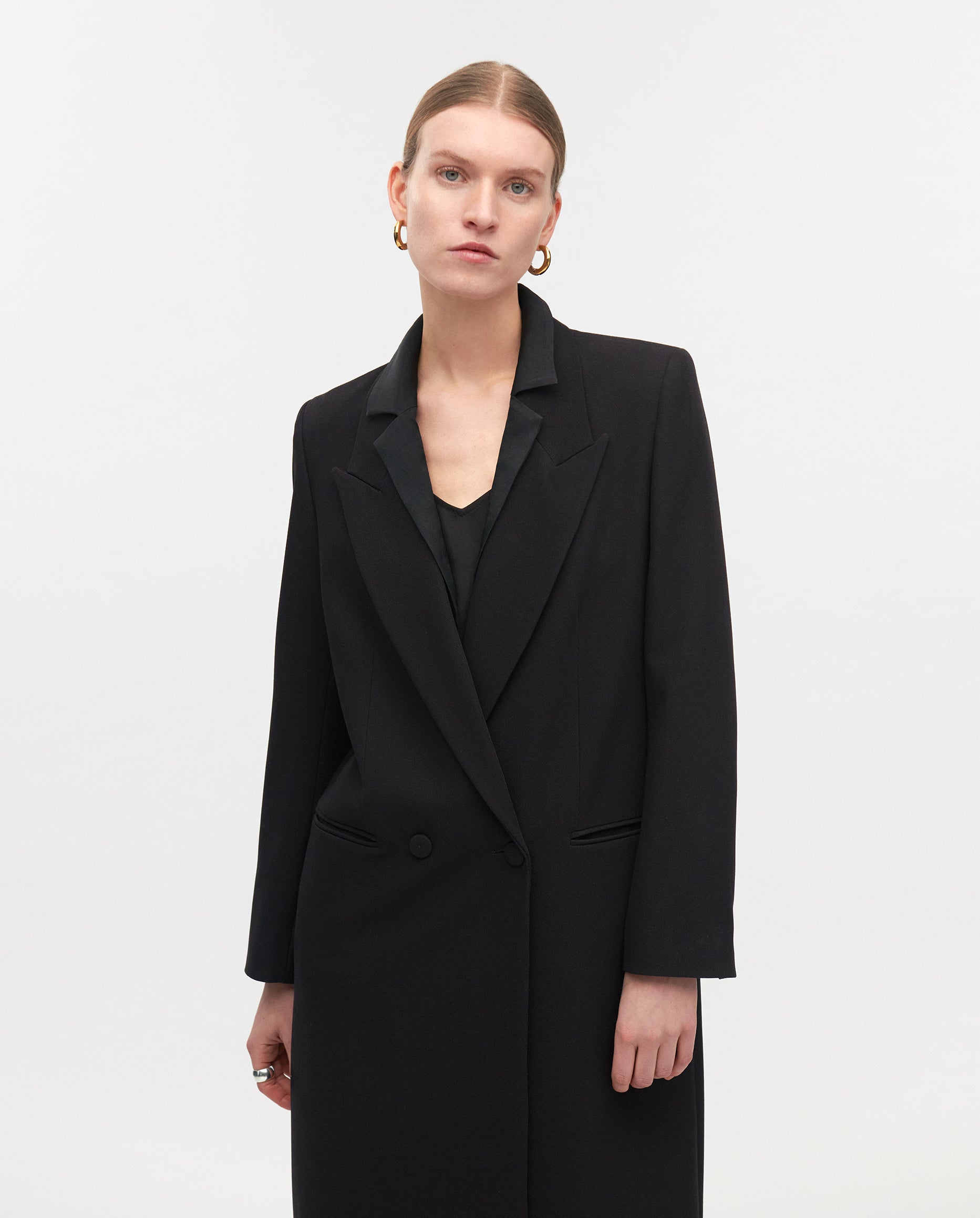 Woman in the IVY OAK JILL ANN Coat, gold hoop earrings, and slicked-back hair against a plain white background—effortlessly showcasing transformable style.