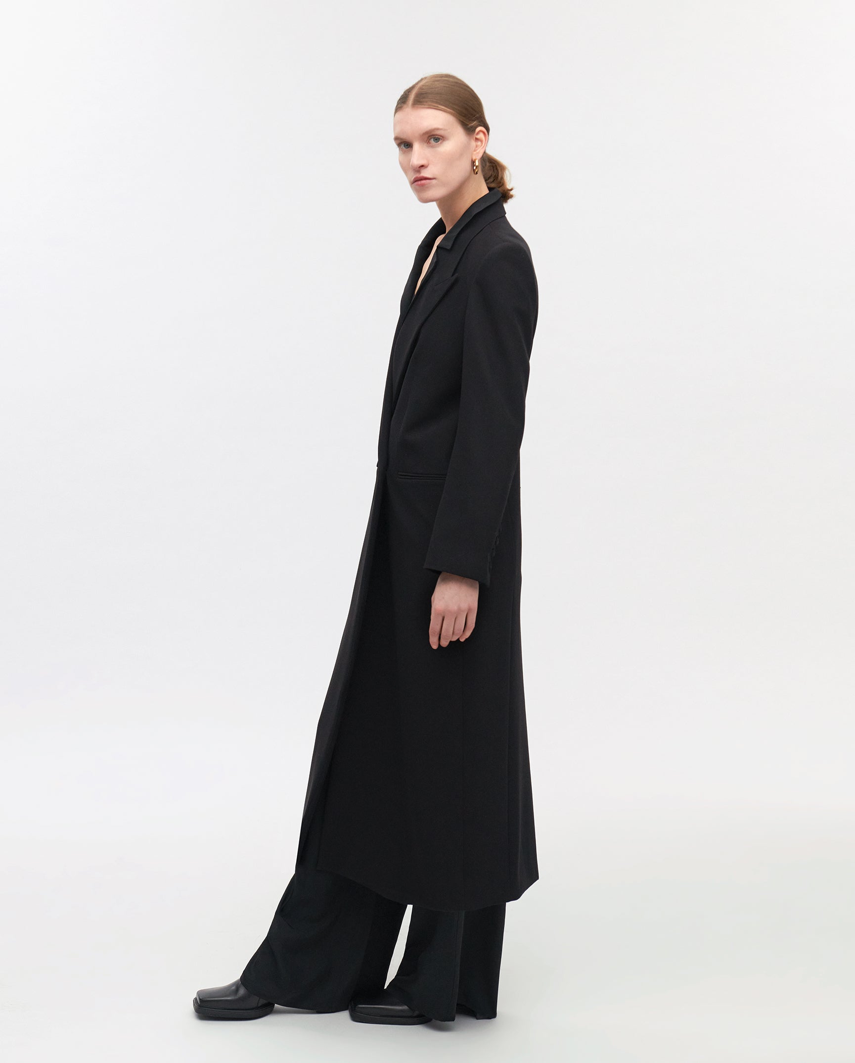 A person stands sideways against a plain background, wearing the IVY OAK JILL ANN Coat in matte fabric, paired with black pants and shoes.