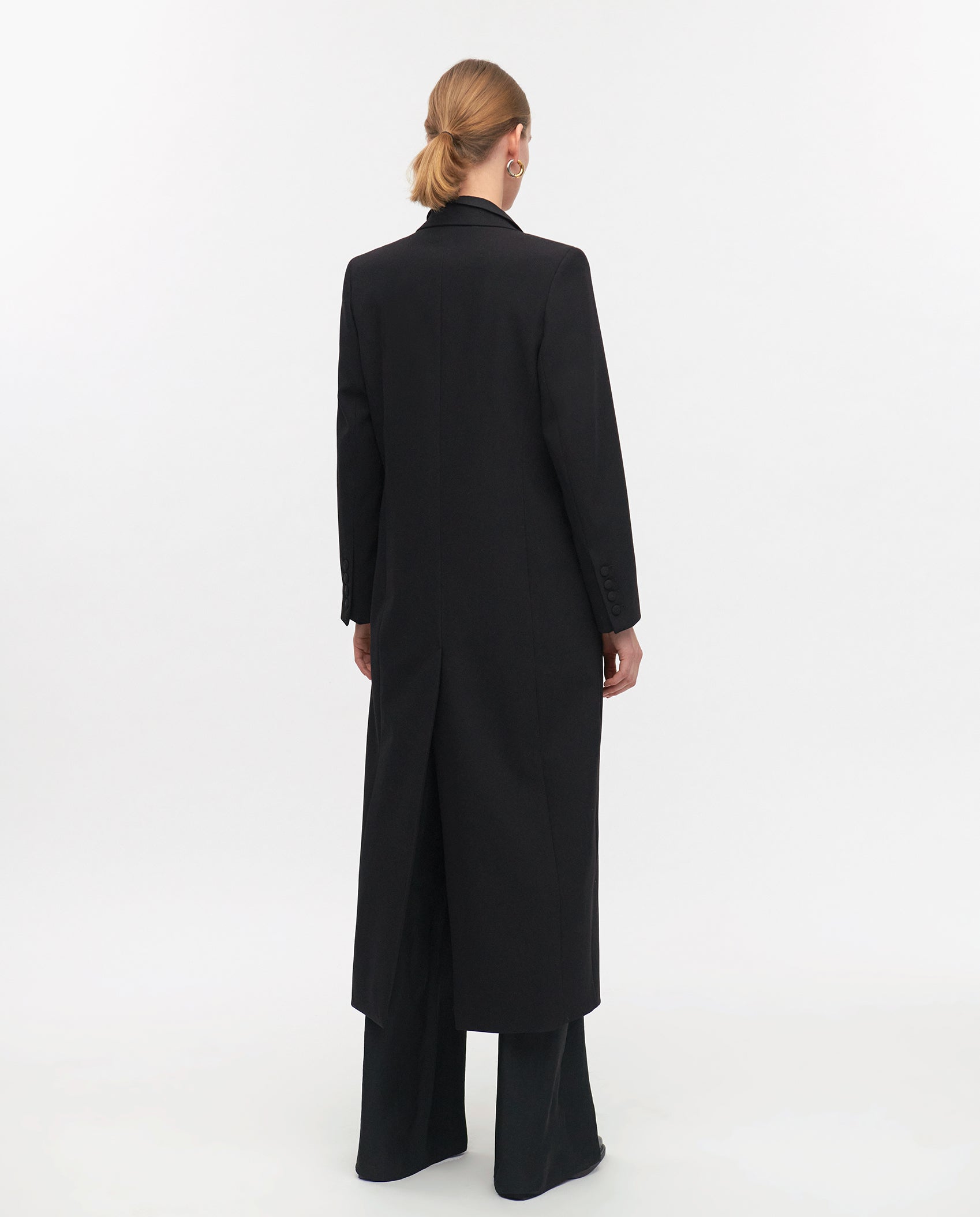 A woman with blonde hair in a bun, facing away from the camera, wears the IVY OAK JILL ANN Coat in matt fabric over black pants.