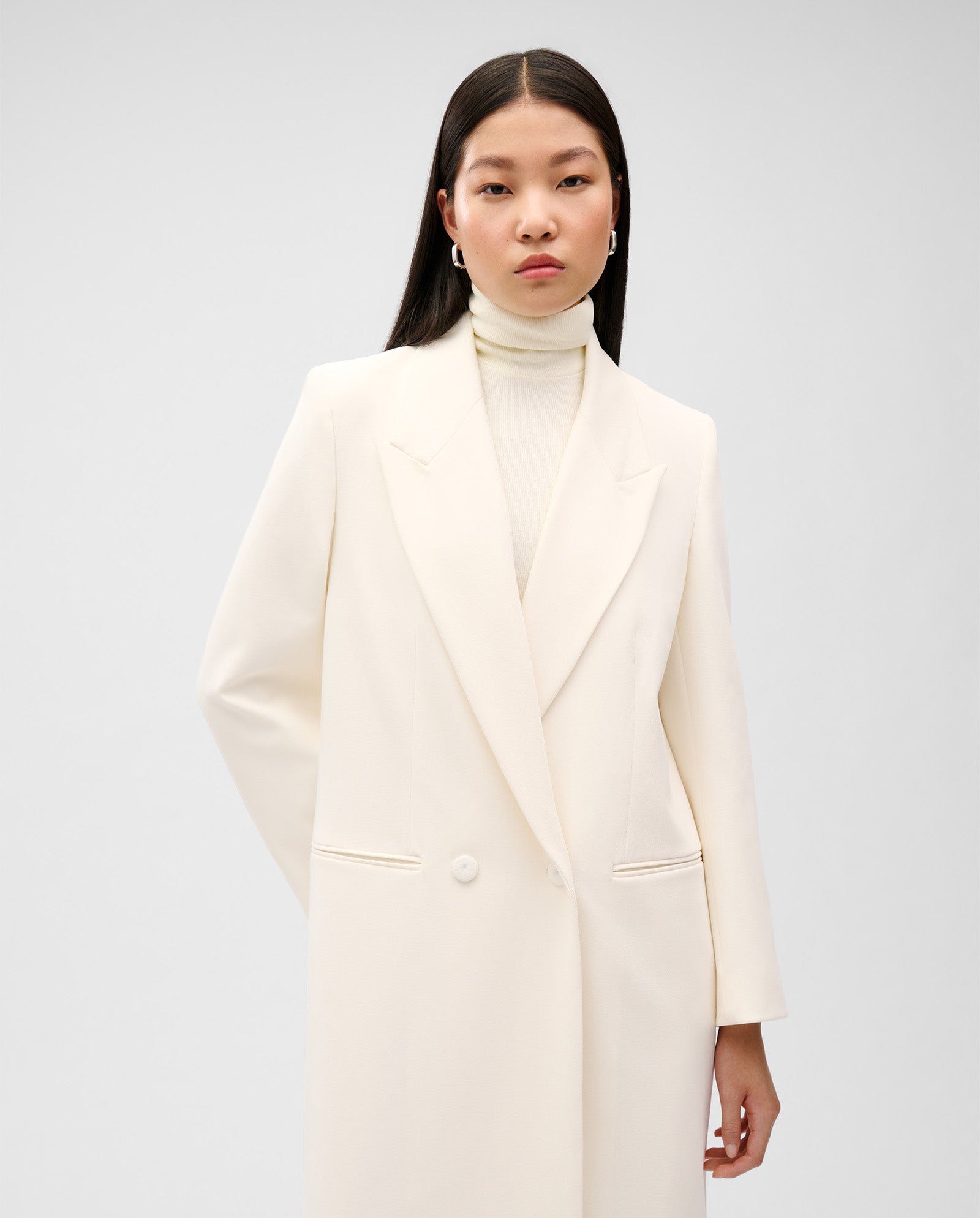 A woman wears the IVY OAK JIL ANN Coat, a long white double-breasted blazer in matt fabric, styled over a white turtleneck against a plain background.