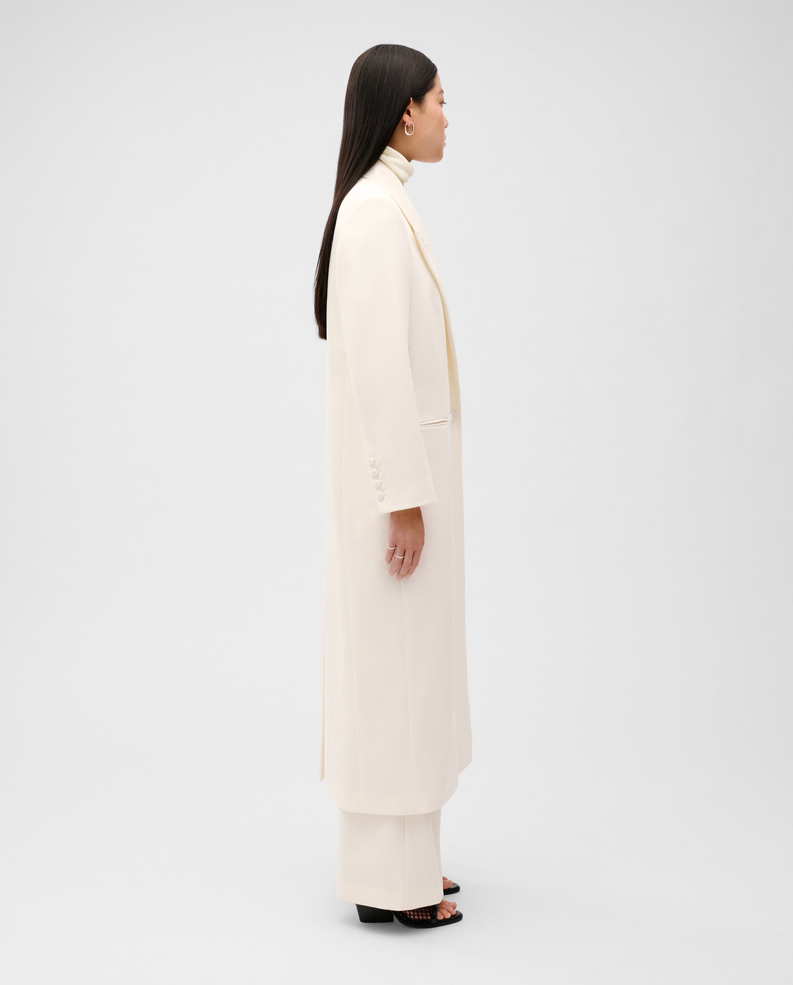 A woman stands sideways against a plain light background wearing the IVY OAK JIL ANN Coat with matching off-white pants.