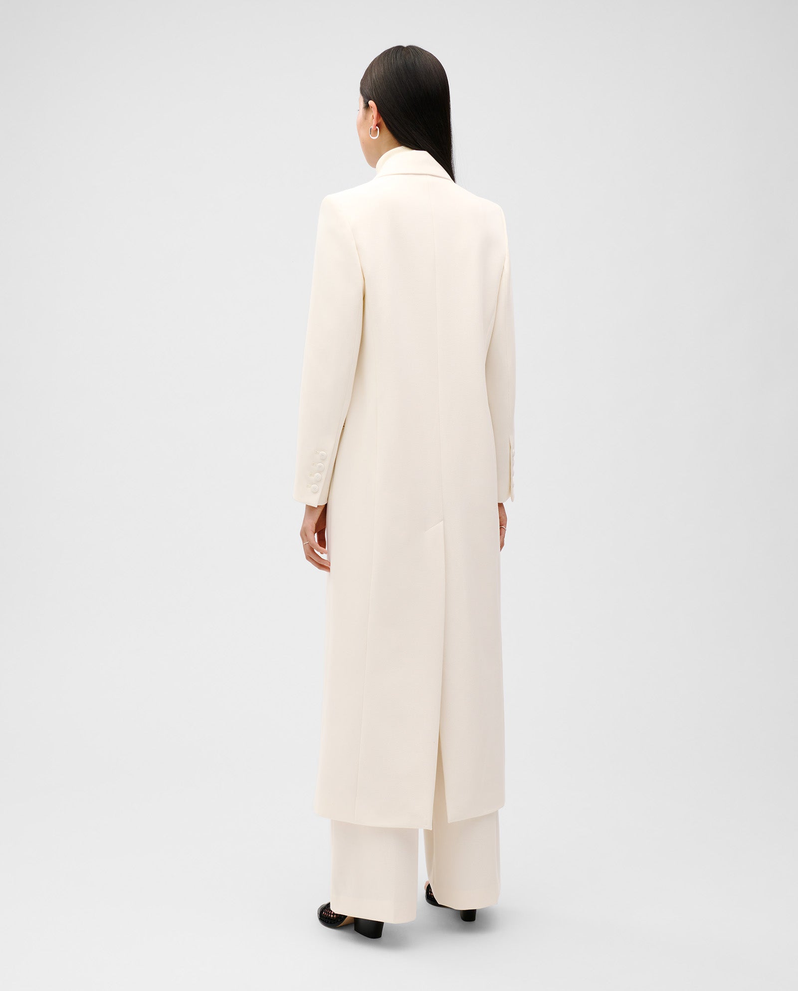 Person wearing the IVY OAK JIL ANN Coat, a long, cream-colored blazer crafted from matte fabric, paired with wide-leg pants and standing with their back to the camera.