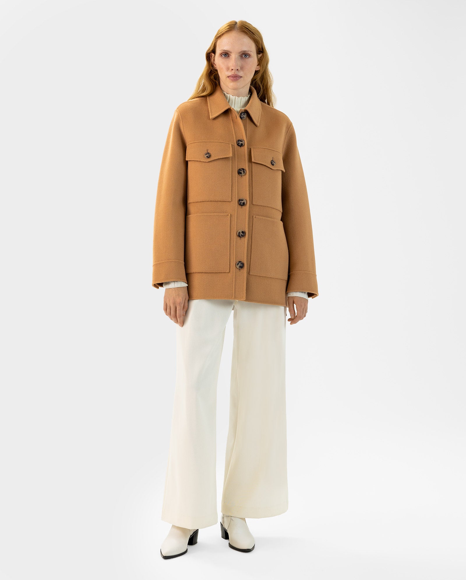 A woman stands against a plain background wearing the IVY OAK JUNO Jacket in tan wool with patched pockets, paired with white wide-leg pants and white boots.
