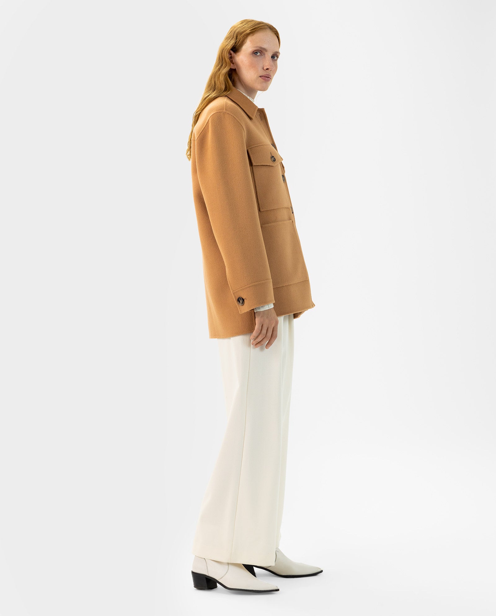 A person wears the IVY OAK JUNO camel wool jacket with patched pockets, white wide-leg pants, and white boots, standing against a plain white background.