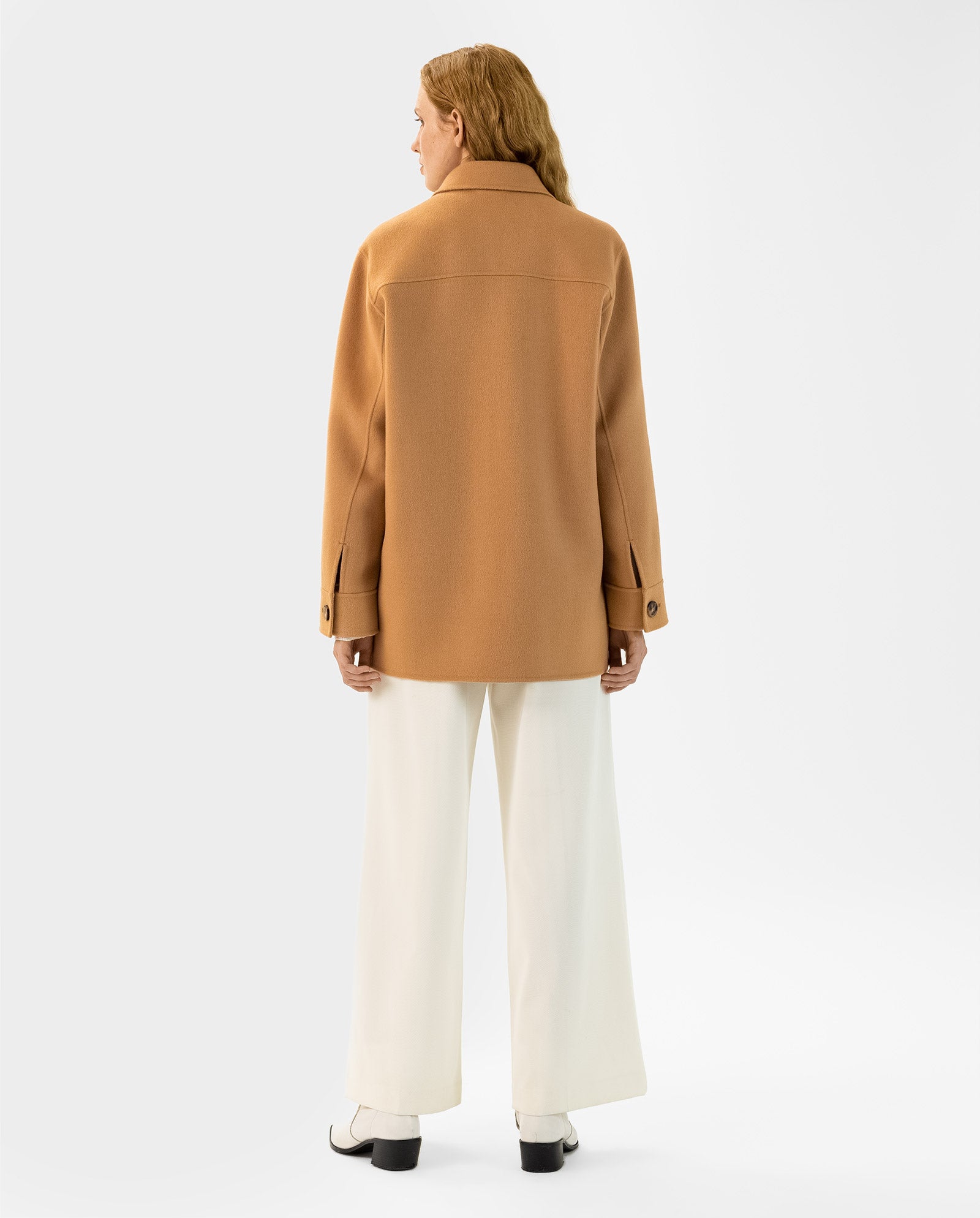 A woman stands facing away, wearing the camel-colored IVY OAK JUNO Jacket with patched pockets, paired with white wide-leg pants and white shoes.