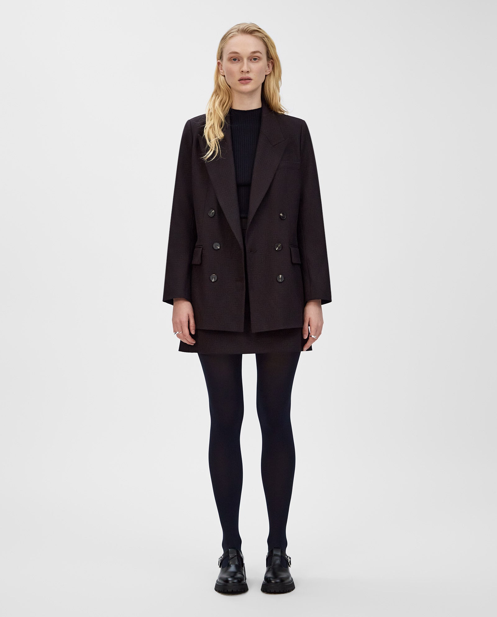 A woman wears the IVY OAK JULIETTE ROSE double-breasted black jacket with a skirt, tights, and shoes, standing against a plain white background.