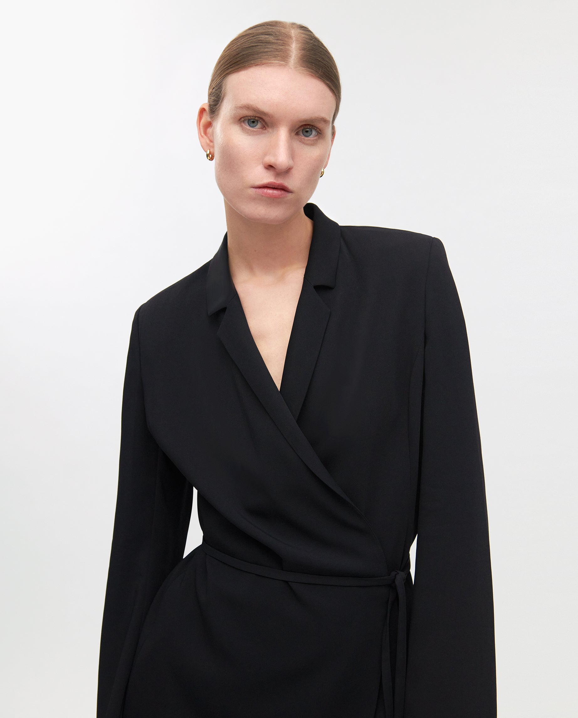 A woman with slicked-back hair wears the black JANIKA Dress by IVY OAK, made from a viscose mix, standing against a plain white background.