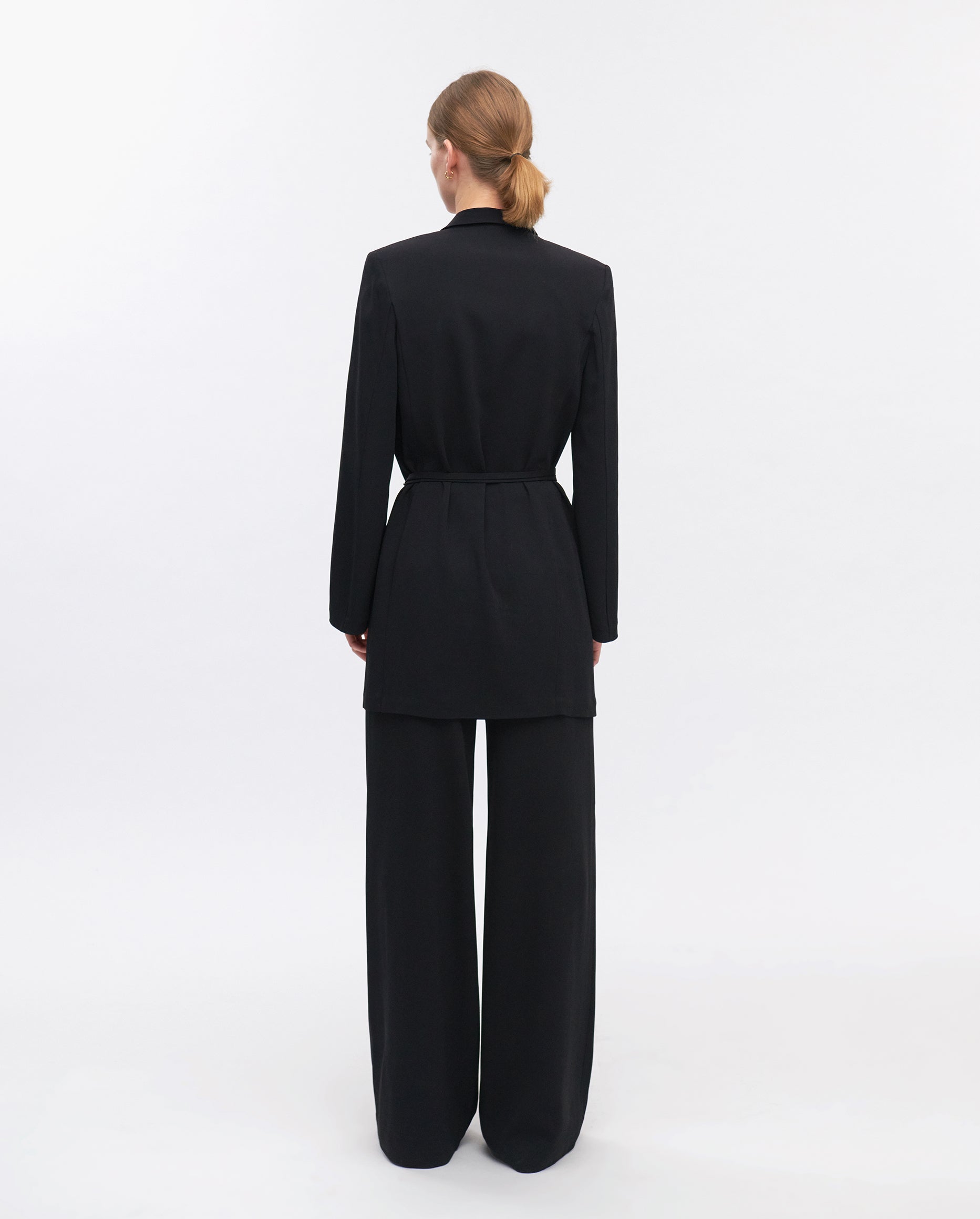 A woman stands facing away against a white background, wearing the JANIKA Dress from IVY OAK in körpernahe Passform and wide-leg black trousers.