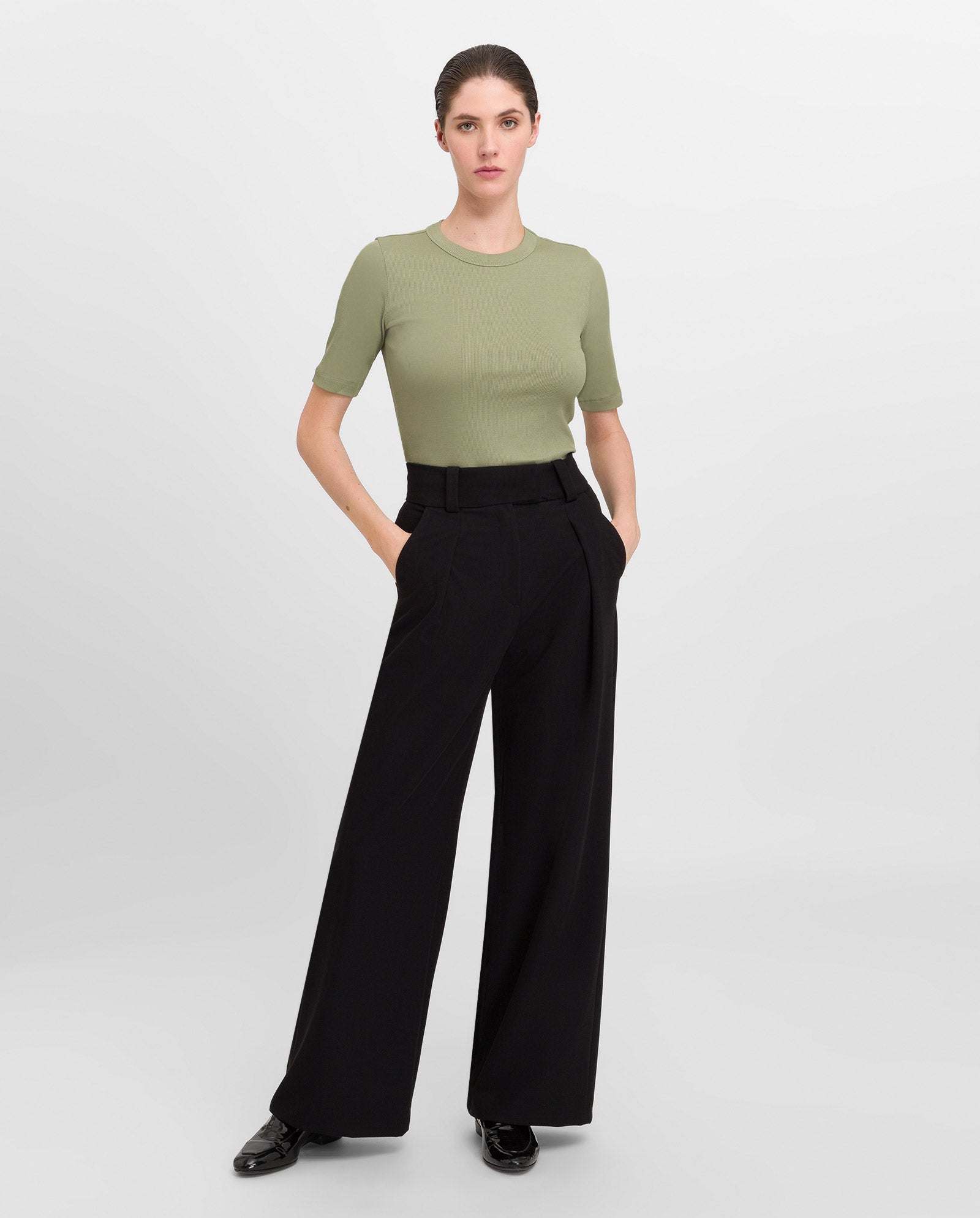 A woman wears the IVY OAK KRISTIN Rib T-Shirt in olive green, made from organic cotton, paired with black wide-leg pants with stretch, standing with her hands in her pockets.