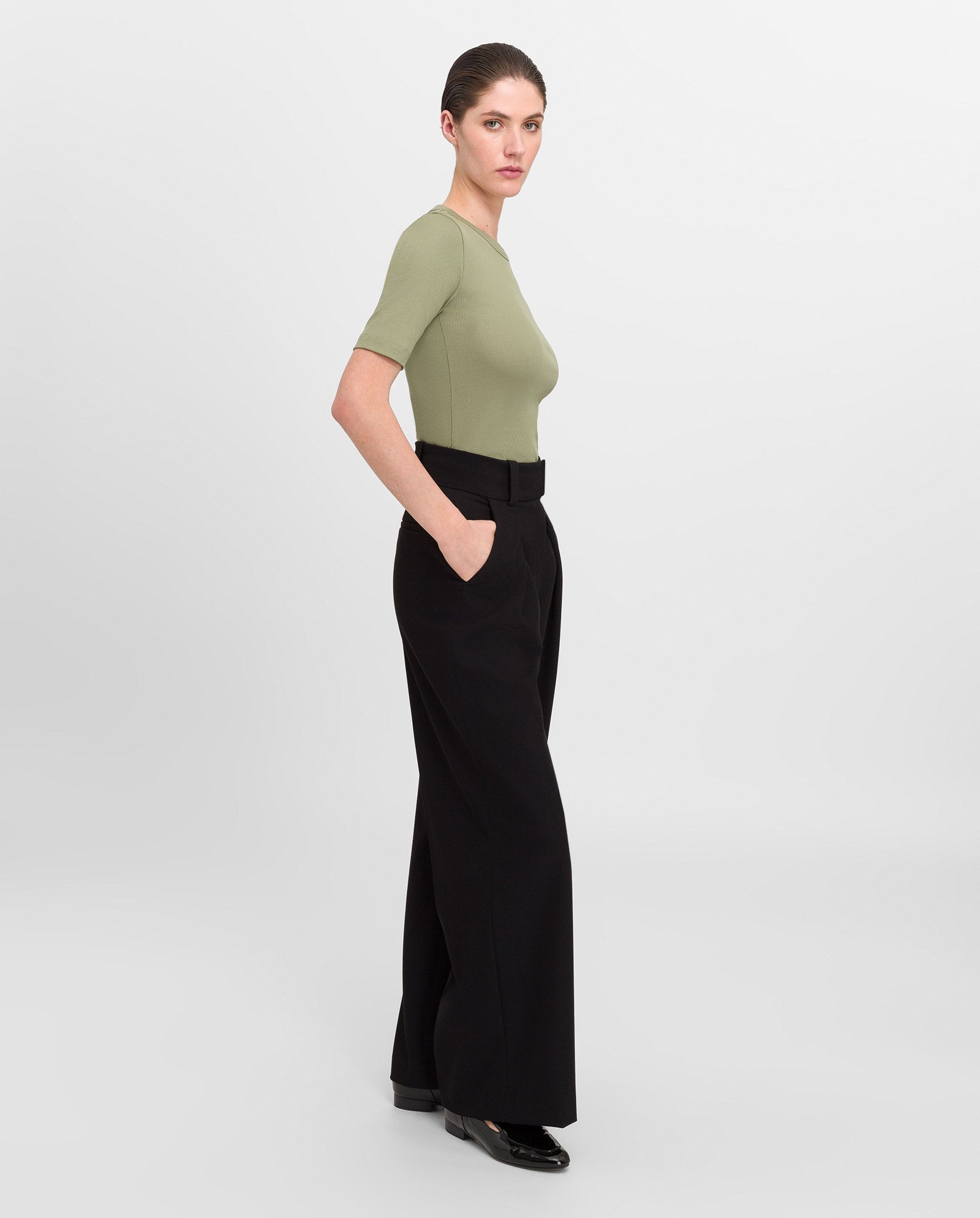 A woman in the IVY OAK KRISTIN Rib T-Shirt—an olive green, fitted top made from organic cotton—and black wide-leg pants stands sideways with her hands in her pockets.