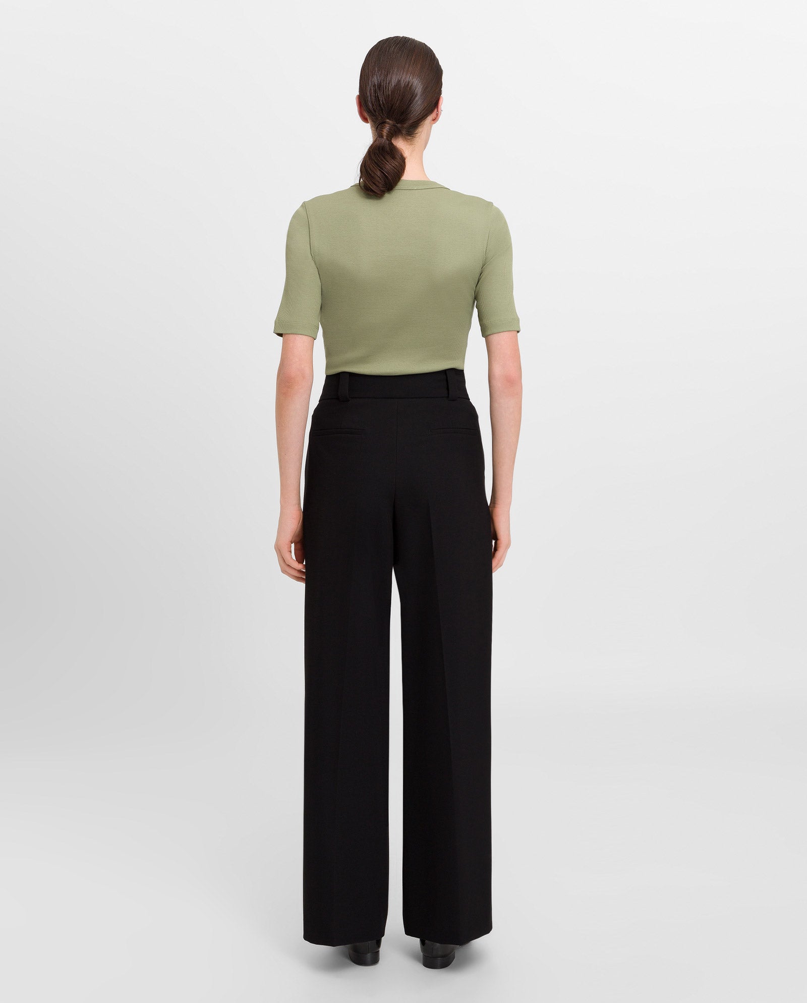 A woman stands with her back to the camera, wearing the IVY OAK KRISTIN Rib T-Shirt in green organic cotton and black wide-leg pants.