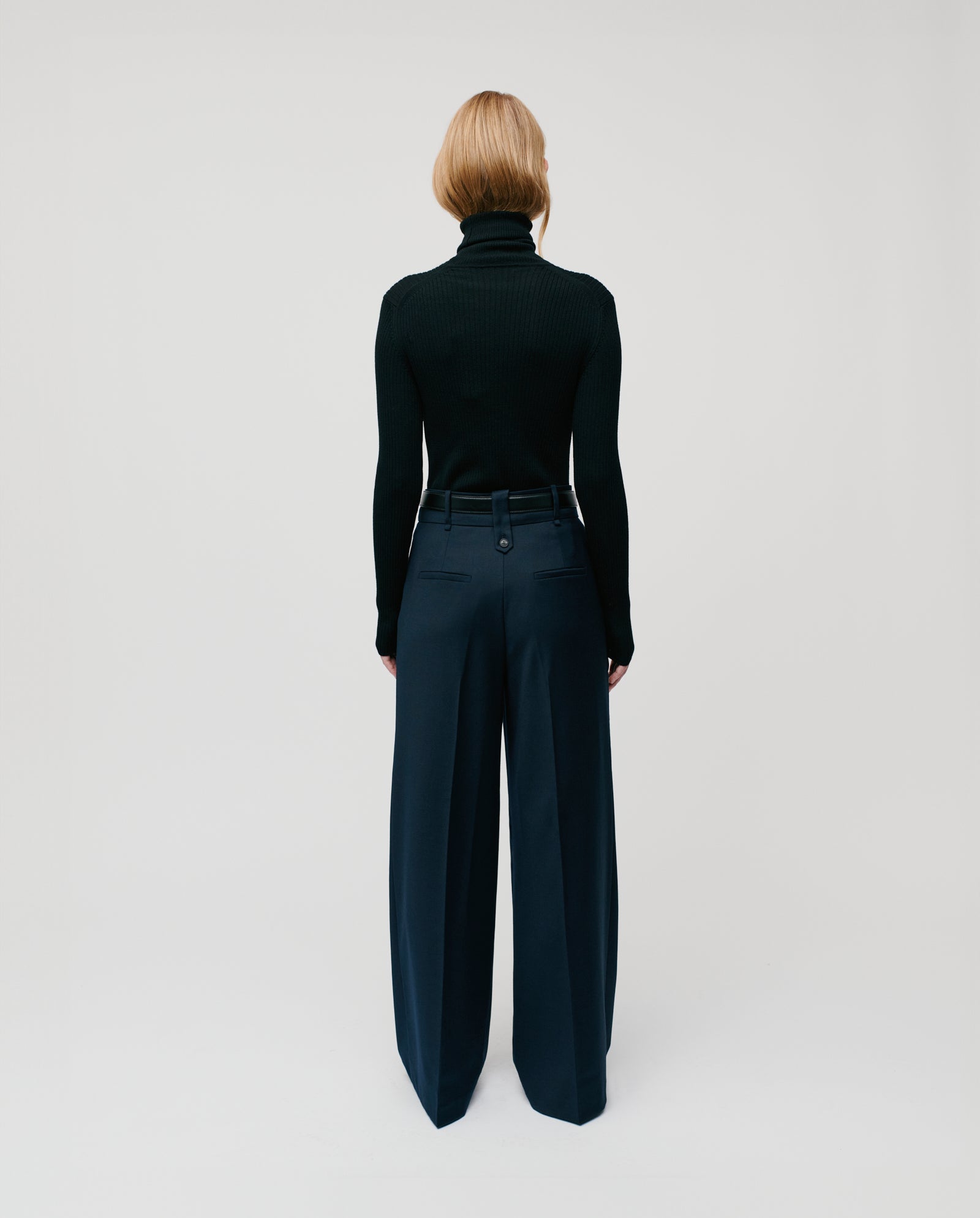 A person with blonde hair, wearing a black KARO Jumper by IVY OAK and navy wide-leg pants, stands facing away.