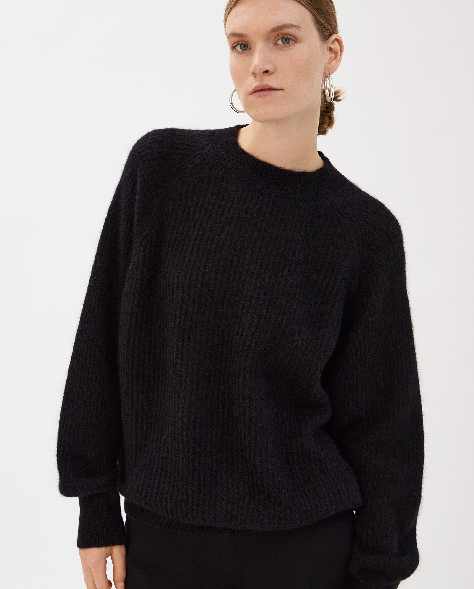 Woman in a loose-fitting black KIRSTEN Jumper by IVY OAK with raglan sleeves and black pants, standing against a plain white background.