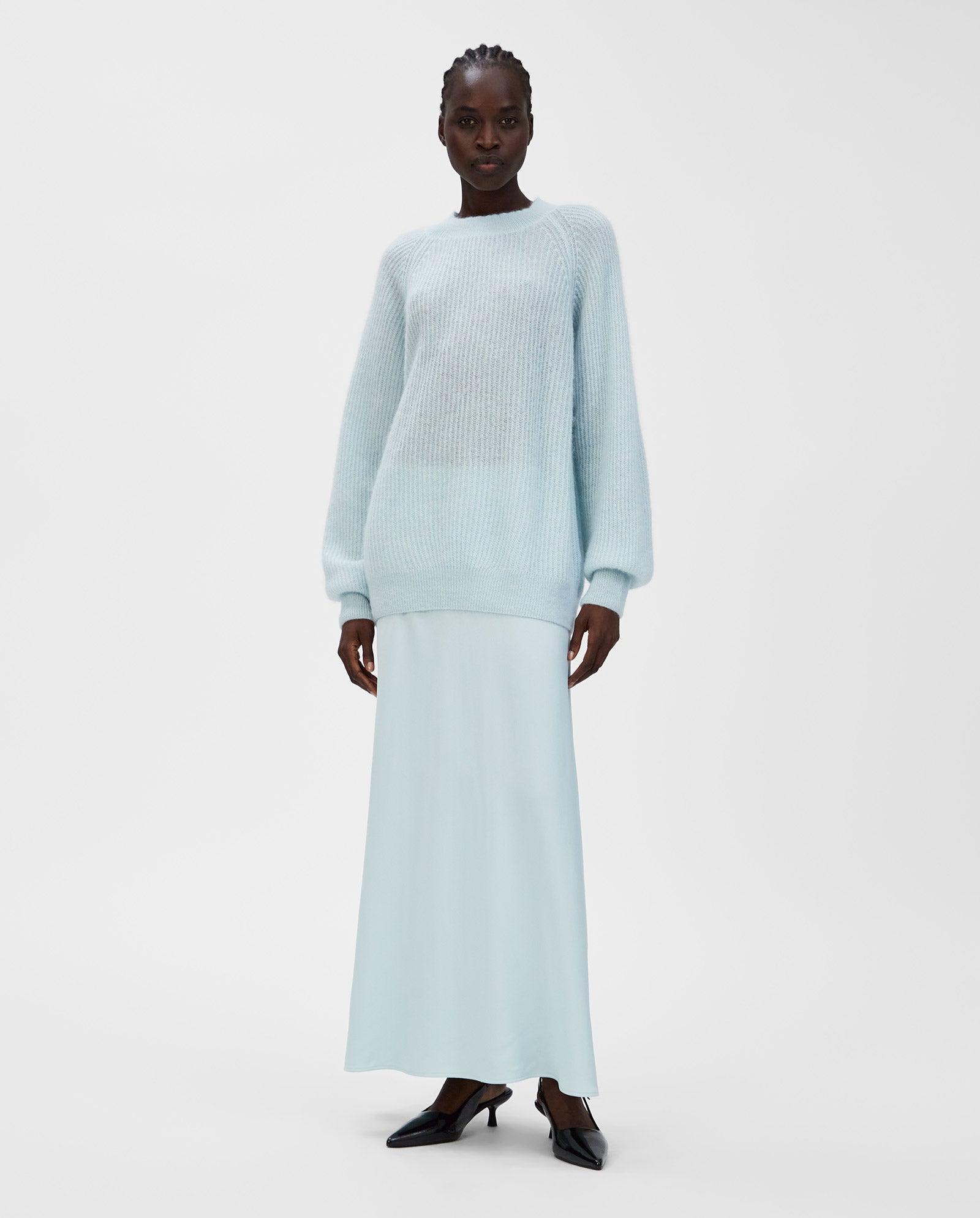 A woman in a light blue KIRSTEN Jumper by IVY OAK with raglan sleeves, paired with a matching long skirt and black heels, stands against a white background.