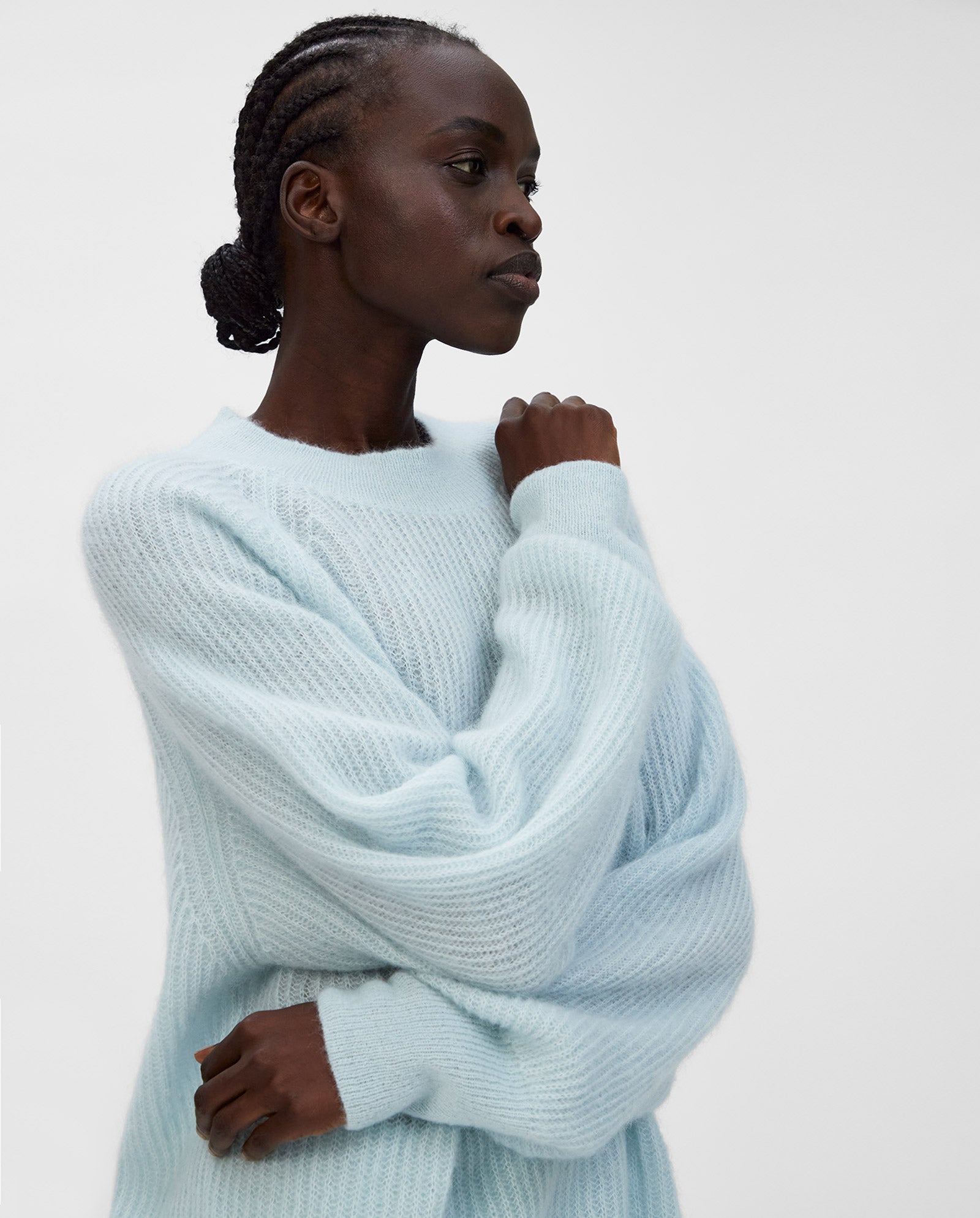 A woman in a light blue, cozy KIRSTEN Jumper by IVY OAK with raglan sleeves stands with one arm raised against a plain, light background.
