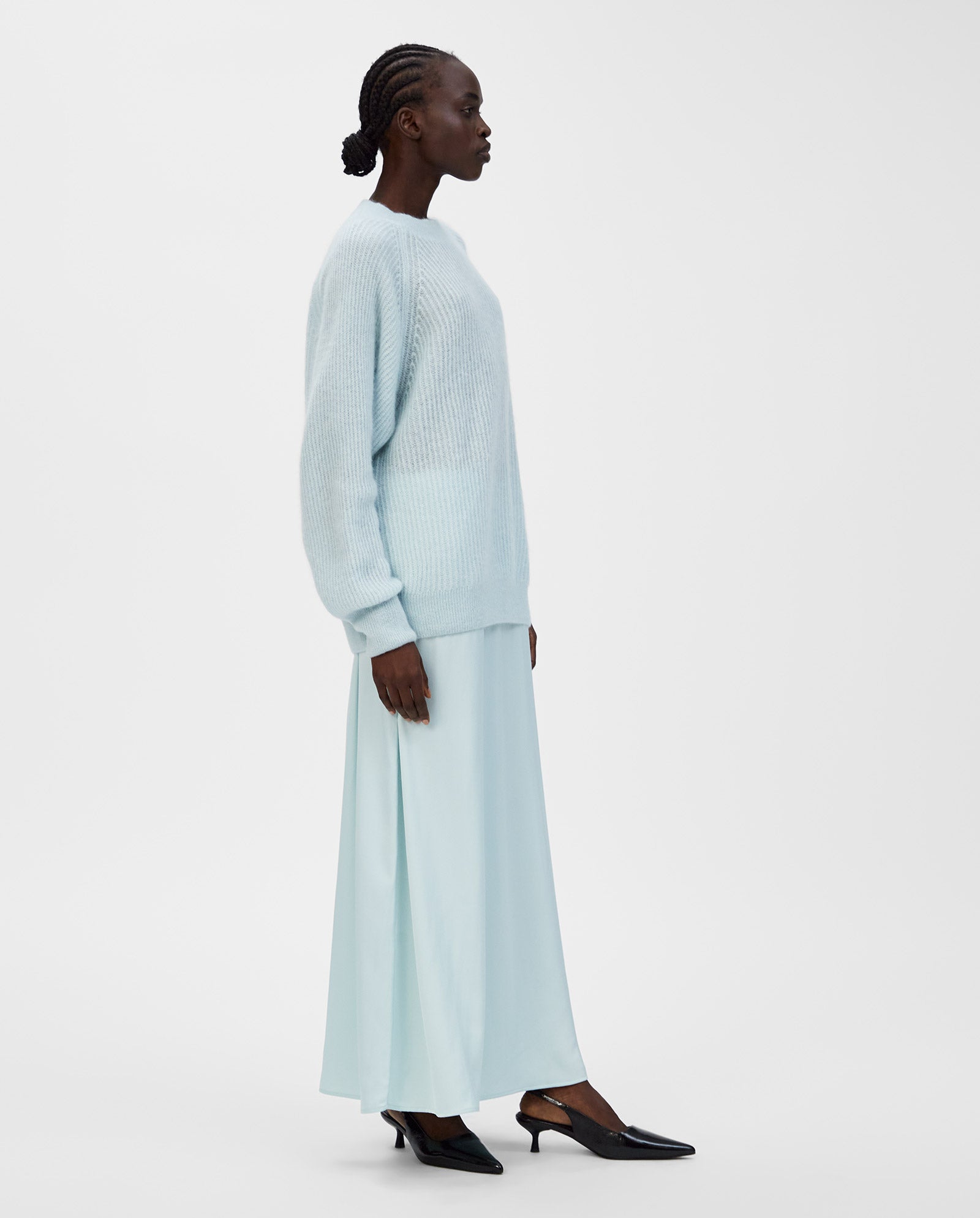A woman in a light blue IVY OAK KIRSTEN Jumper with raglan sleeves and a skirt, paired with black heels, stands in profile against a white background.